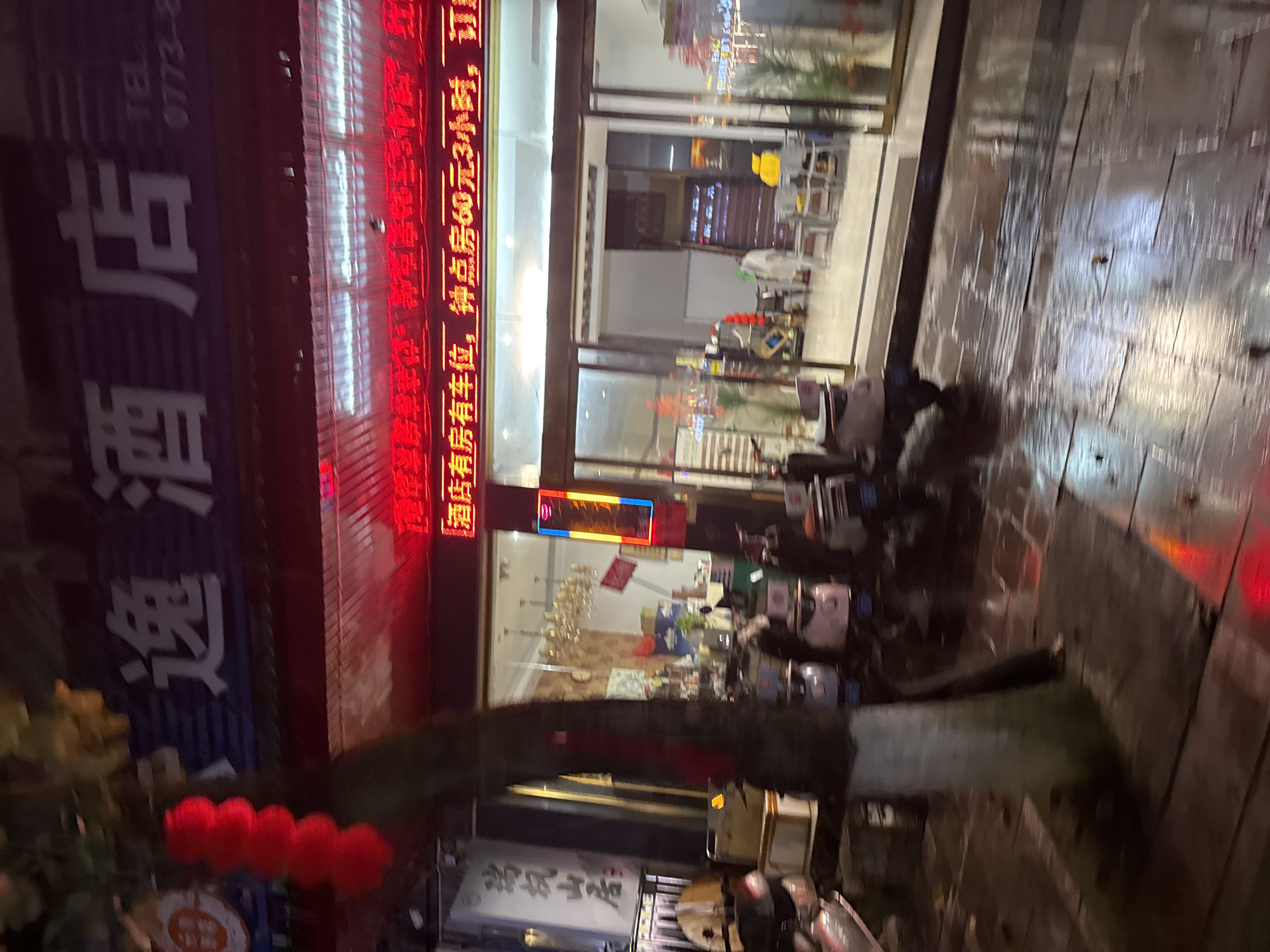 The image shows a street scene in Guilin, China at night. The focus is on a building with a sign indicating "逸酒店" (Yi Hotel) in vertical, lit characters. Above it, there is another illuminated sign displaying information in red Chinese characters, including details about hourly room rates and parking availability. The street appears wet, possibly due to recent rain, with reflections on the pavement. There are several parked motorbikes along the sidewalk. A tree with red lanterns is visible in the foreground.