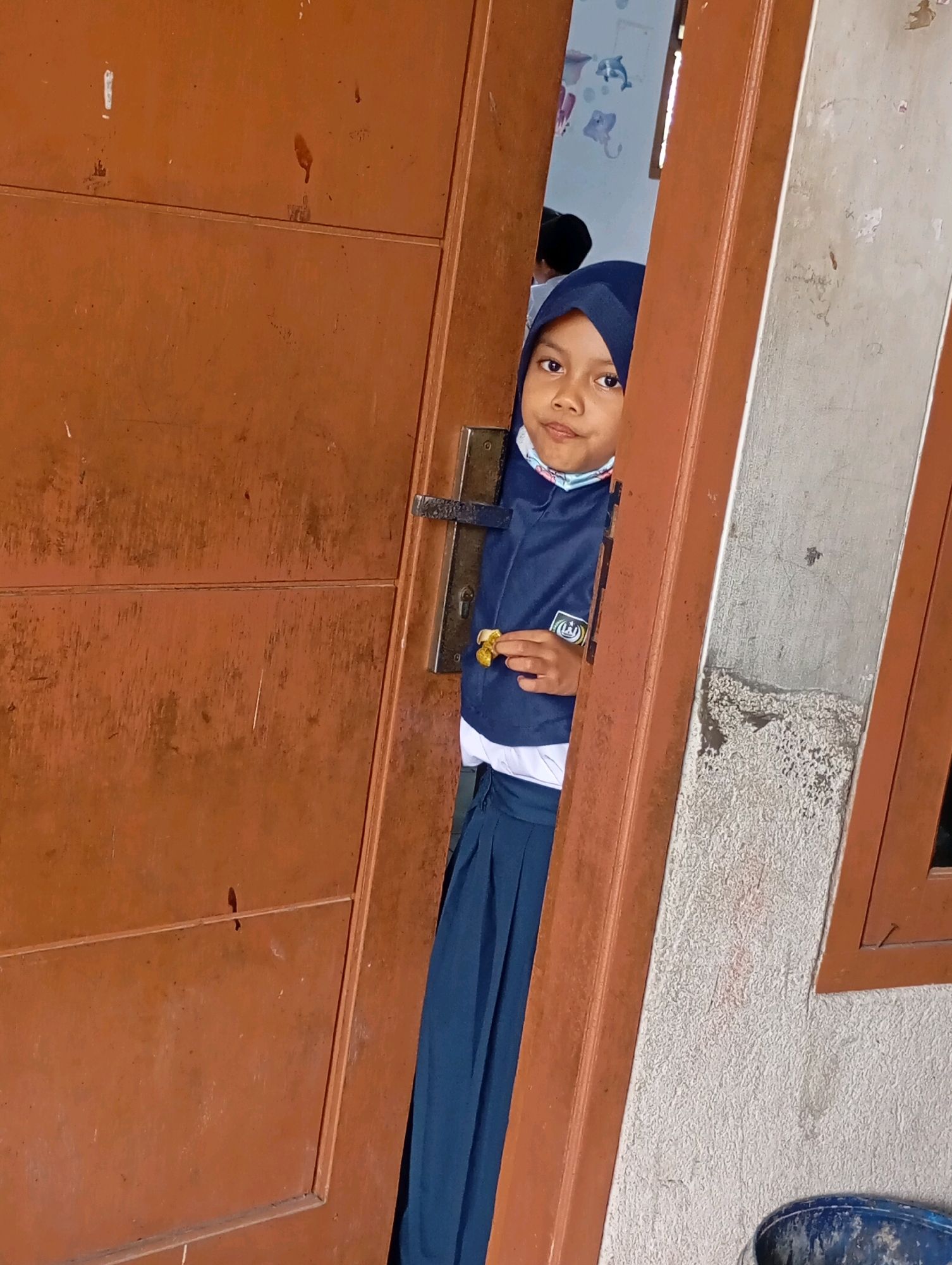 A young girl wearing a navy blue hijab, a white shirt, and a navy blue skirt stands peeking through an open wooden door. She is holding a small yellow object in her right hand. The door is dark brown and appears to be made of wood. To the right of the door is a textured, light-colored wall with some scuff marks. In the background, inside what appears to be a room, a portion of a wall with colorful drawings of sea creatures is visible. The scene suggests a residential setting in Babelan, Indonesia, possibly a home or school.