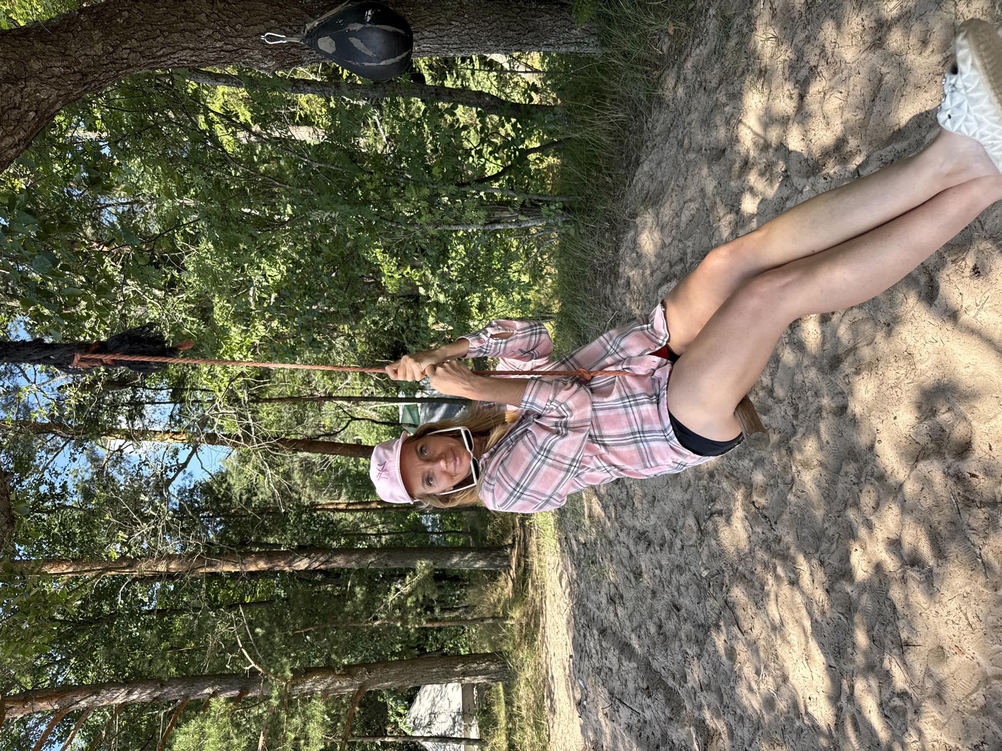 A woman with blonde hair wears a pink plaid shirt, black shorts, and a pink baseball cap while sitting on a swing in a sandy, wooded area. She is holding onto the rope of the swing with both hands, her sunglasses are perched on her head. The background shows trees, some with tents visible behind them. The ground is covered in sand with dappled sunlight and shadows. A punching bag is attached to a tree to the right of the woman. The location is identified as Ozerki (Krasnodolinskaya), Russia.