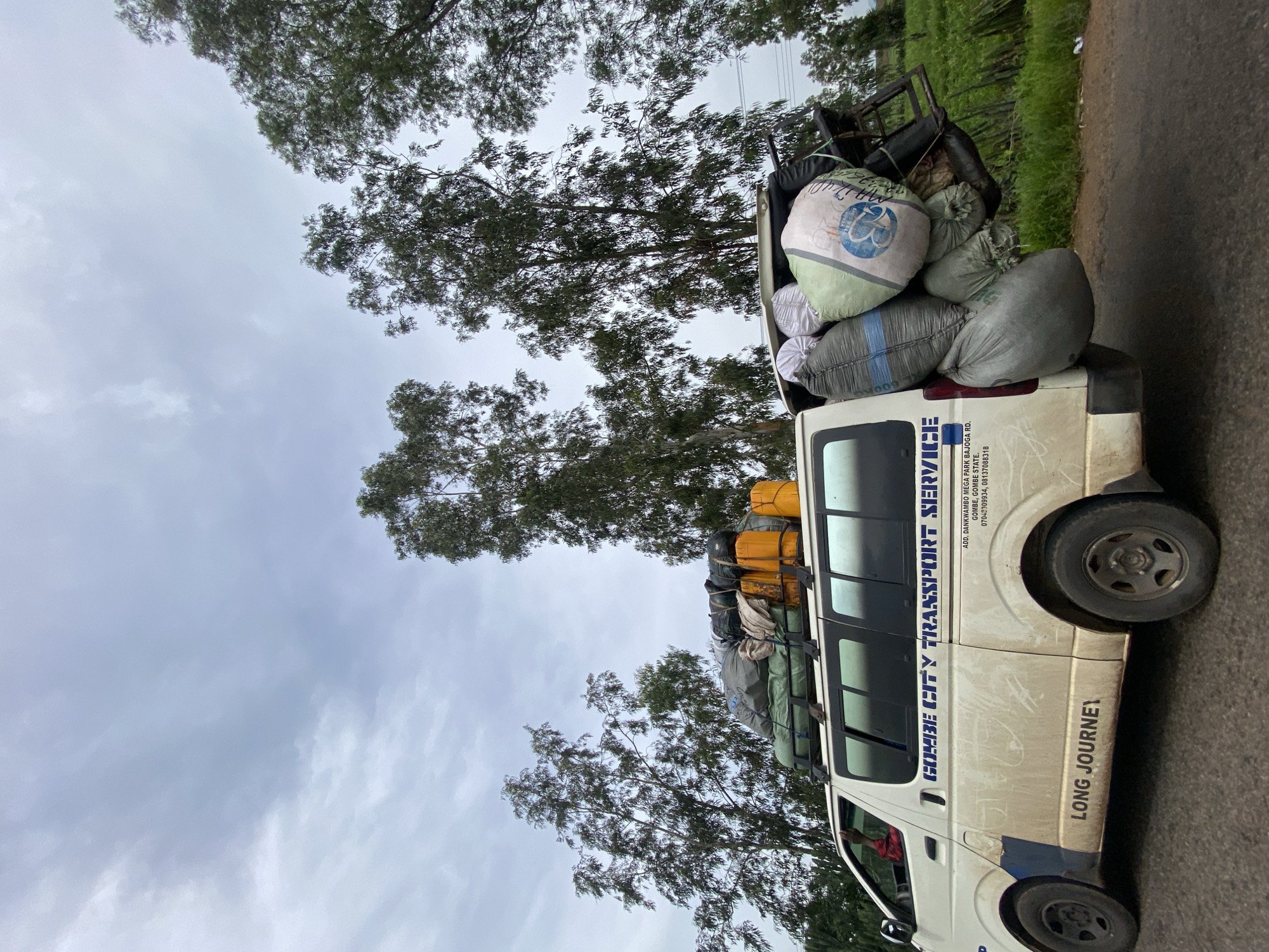 A white van labeled "GOMBE CITY TRANSPORT SERVICE" is parked on a road. The van is heavily laden with luggage and cargo, both on its roof and attached to its rear. The roof rack is filled with bags, yellow canisters, and other assorted items. A metal rack at the back of the van is also overflowing with large sacks. The van's side has the words "LONG JOURNEY" written on it. The scene appears to be in a rural or semi-rural area with tall trees lining the road. The sky is overcast, suggesting a cloudy day. The image does not provide explicit information about the city of Hoss, Nigeria, or any specific location within it.