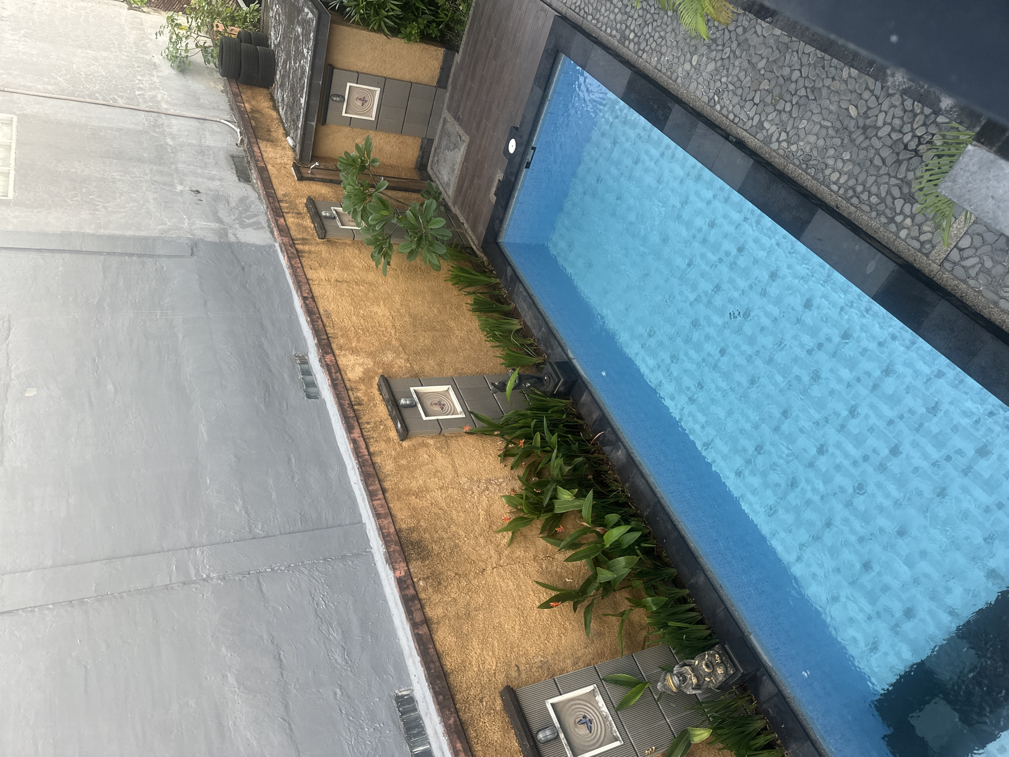 An aerial view of a small, rectangular swimming pool in a courtyard in Kuta, Indonesia. The pool is filled with clear blue water and has a tiled bottom. The pool is surrounded by a dark tiled border and a stone walkway. To the left of the pool, there is a wall with decorative tiles and a small statue. To the right, there is a wooden deck with a small building and some greenery. The background shows a grey wall of a building. The overall scene is one of a private residence or hotel.