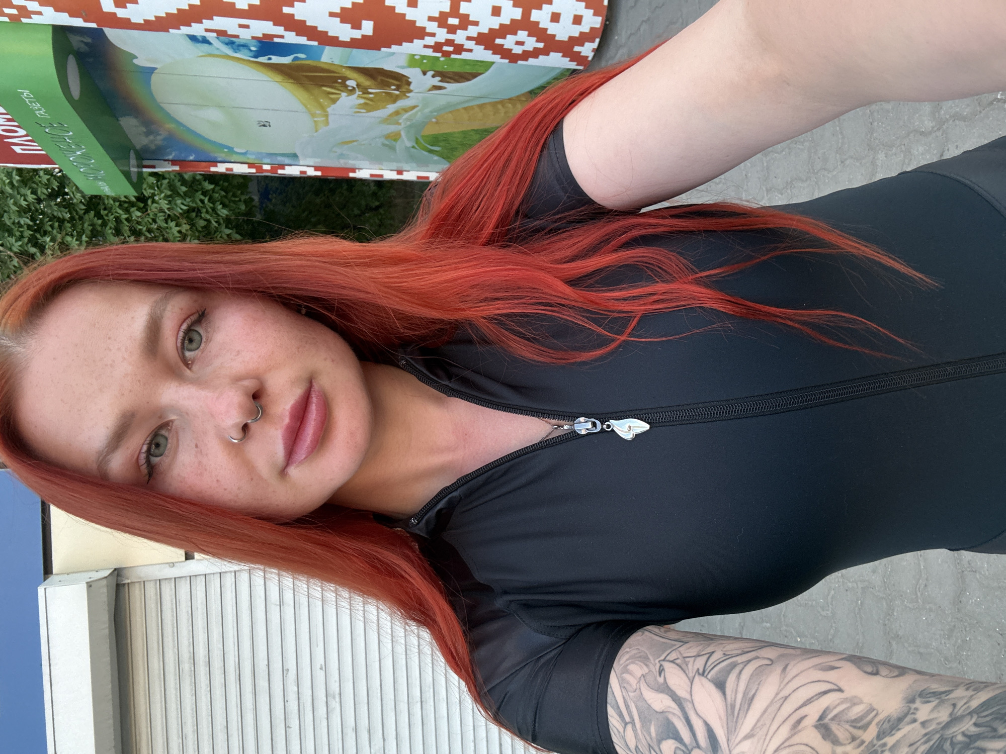 A young woman with red hair and freckles poses in a selfie taken outdoors. She has a nose ring and wears a black top with a zipper. Her left arm, visible in the lower right corner, has a tattoo. The background includes a building with a roller shutter door, a sign featuring an ice cream cone and text in Russian, and some greenery. The image was taken in the city of Rostov-on-Don, Russia.