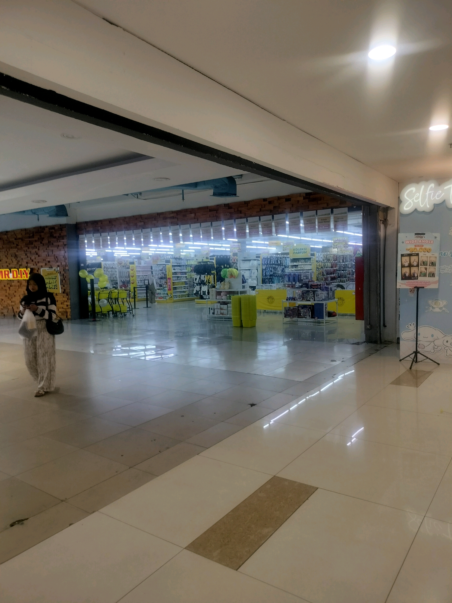 The image captures the interior of a MR. DIY store in Batam, Indonesia, as indicated by the signage. The scene is brightly lit by fluorescent lights reflecting off the polished tiled floor. In the foreground, a woman wearing a hijab and a patterned outfit walks towards the left, holding a plastic bag and a phone. She is looking down at her phone. The entrance to the MR. DIY store is visible in the background, revealing rows of shelves stocked with various products, including what appear to be home improvement and retail items. Yellow promotional balloons and shopping carts are present near the entrance. To the right of the store entrance, a stand with a poster featuring a "Selfie" theme and product advertisements is set up. The overall environment suggests a well-lit and organized retail space within a commercial area.