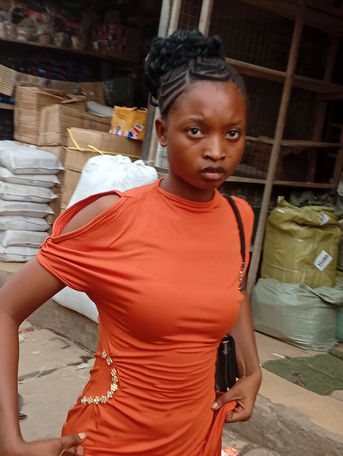 A young woman with intricately braided hair pulled into a bun poses outdoors, wearing a bright orange dress with a shoulder cutout. The dress features a decorative gold embellishment at the waist. She holds the hem of her dress, looking directly at the camera. The background is a bustling marketplace with stacked goods and a wooden shelving structure. The setting suggests a lively, commercial environment.