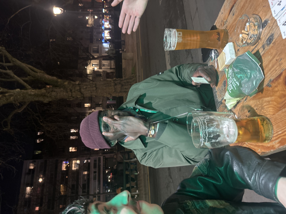 At night, three people sit at a wooden table outside. An African-American man wearing a burgundy beanie and olive green jacket covers his face with his hand, holding playing cards. Beside him, a person in a green jacket is partially visible. Another hand hovers in the upper portion of the frame. The table holds a full beer glass, an empty beer glass, an ashtray filled with cigarette butts, a pack of cigarettes, and a snack bag. Buildings with lit windows appear in the background.