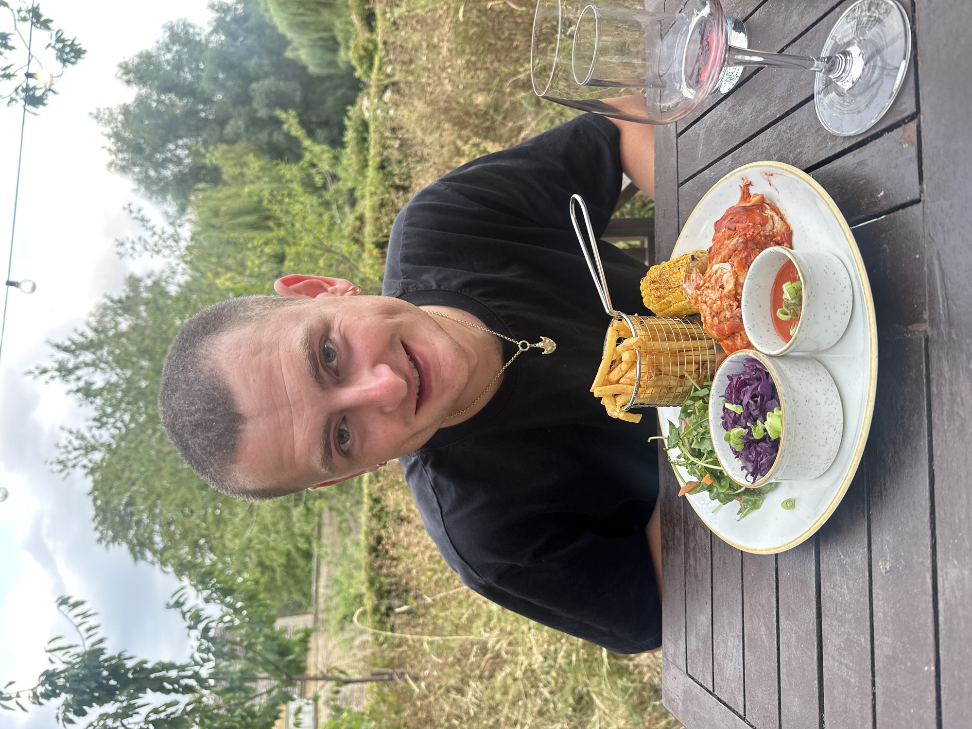 A young man with a shaved head and a black t-shirt smiles at the camera while sitting at a wooden table outdoors. He has a gold necklace with a pendant and a hoop earring in his left ear. In front of him is a plate of food that includes french fries in a metal basket, a piece of corn on the cob, a salad with red cabbage, and what appears to be a piece of meat covered in a red sauce. There is also a small dish of red sauce with green onions on top. To the right of the plate are two wine glasses, one of which contains red wine. The background is blurred and shows trees and foliage, suggesting a garden or outdoor dining area. The lighting indicates it is daytime.