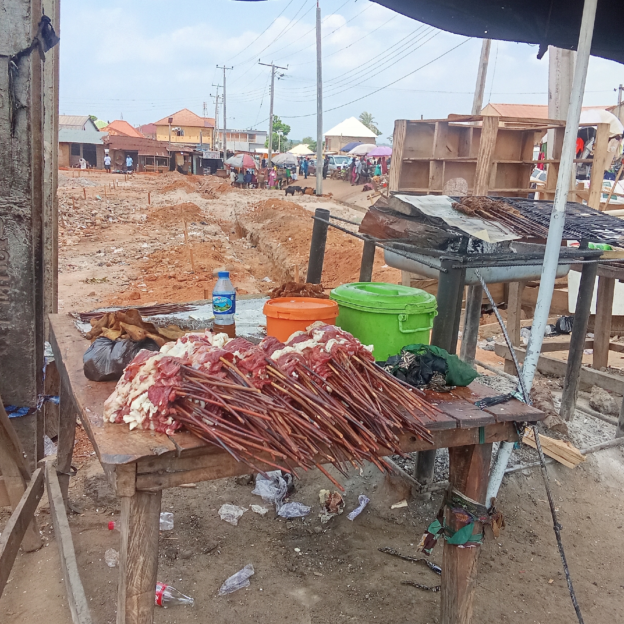The image depicts an outdoor food vending scene, potentially in Akwanga, Nigeria. A wooden table laden with skewers of raw meat dominates the foreground. Beside the meat are a bottle of water, a black plastic bag, and two colorful containers: one orange and one green. Behind the table is a charcoal grill with already cooked skewers. The background reveals a street scene, with several people clustered in small groups, some under umbrellas, in an open area. Dirt and uneven ground indicate potential construction or road work. Buildings and utility poles are visible in the background. Debris is scattered on the ground under the table.
