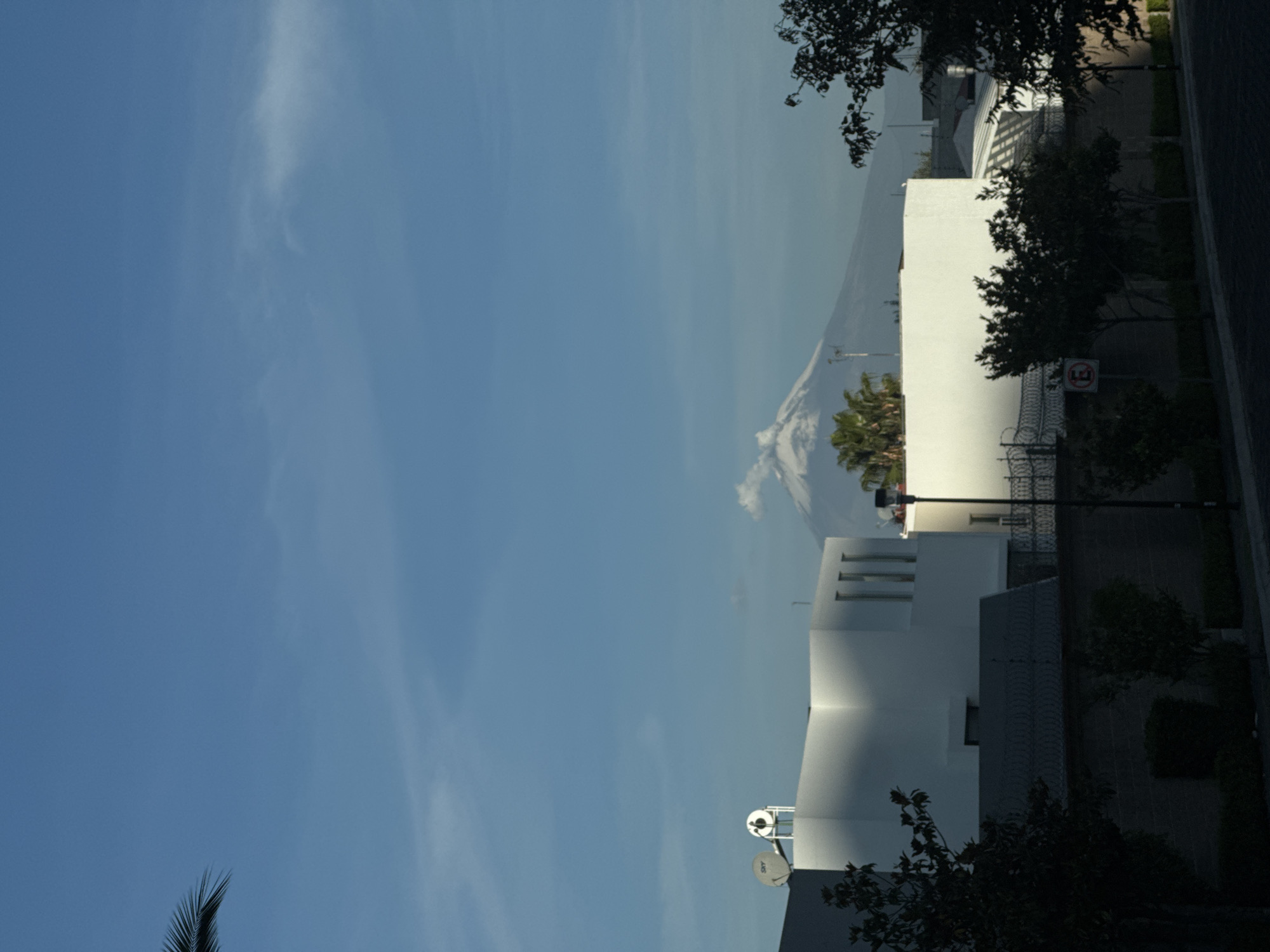 The image depicts a partly cloudy sky above a white, multi-level building in San Bernardino Tlaxcalancingo, Mexico. The top of a volcano is visible in the background, partially obscured by clouds. A palm tree and other vegetation are also visible in the scene. A street with trees and streetlights runs along the side of the building. A satellite dish is mounted on the building.