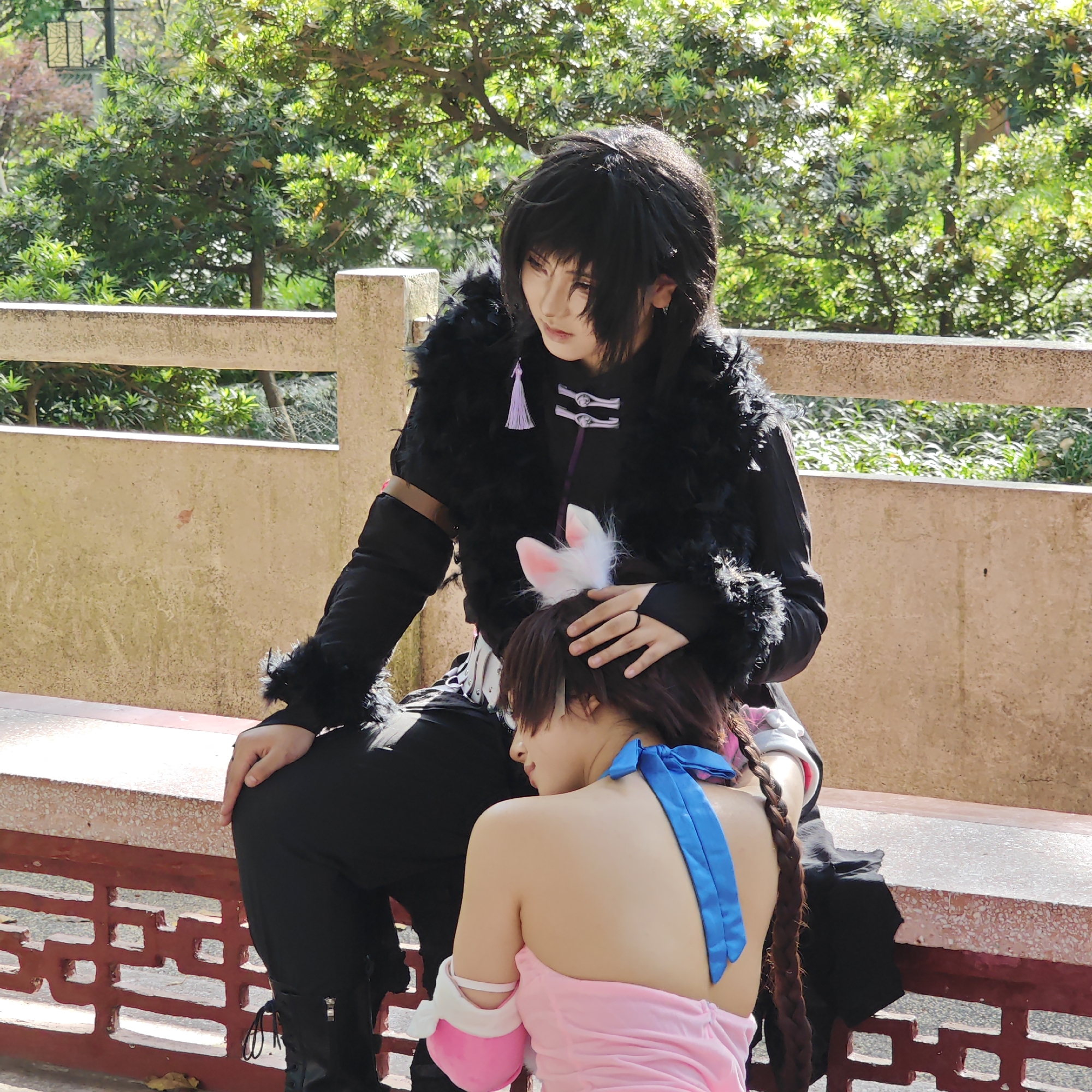 This image depicts two individuals in cosplay, possibly within a park or garden setting in Yuzhong District, China, under bright daylight.
One person is seated on a stone bench, wearing an elaborate black costume with a black faux fur collar and cuffs. They have a black wig with bangs, styled to frame their face, and light-toned stage makeup. Their outfit includes a black top with distinctive silver-colored closures and a small light purple tassel hanging from the collar. They wear black pants and boots, and their right hand rests gently on the head of the other individual.
The second person is positioned in front of and leaning against the seated individual, their head resting on the first person's leg or lap. They are dressed in a sleeveless, vibrant pink dress with a striking blue ribbon tied around their neck, forming a bow at the back. White and pink arm cuffs are visible on their forearms. Adorning their head are white and pink cat ears, and a long, dark brown braid cascades down their back.
The pose conveys a moment of tenderness or comfort, suggesting a close bond between the two subjects. The background is filled with lush green trees and foliage, indicating an outdoor, natural environment. A light-colored stone or concrete railing runs horizontally behind the subjects, and part of a textured wall structure is also visible. The sunny weather casts clear shadows. No discernible text is visible in the image.