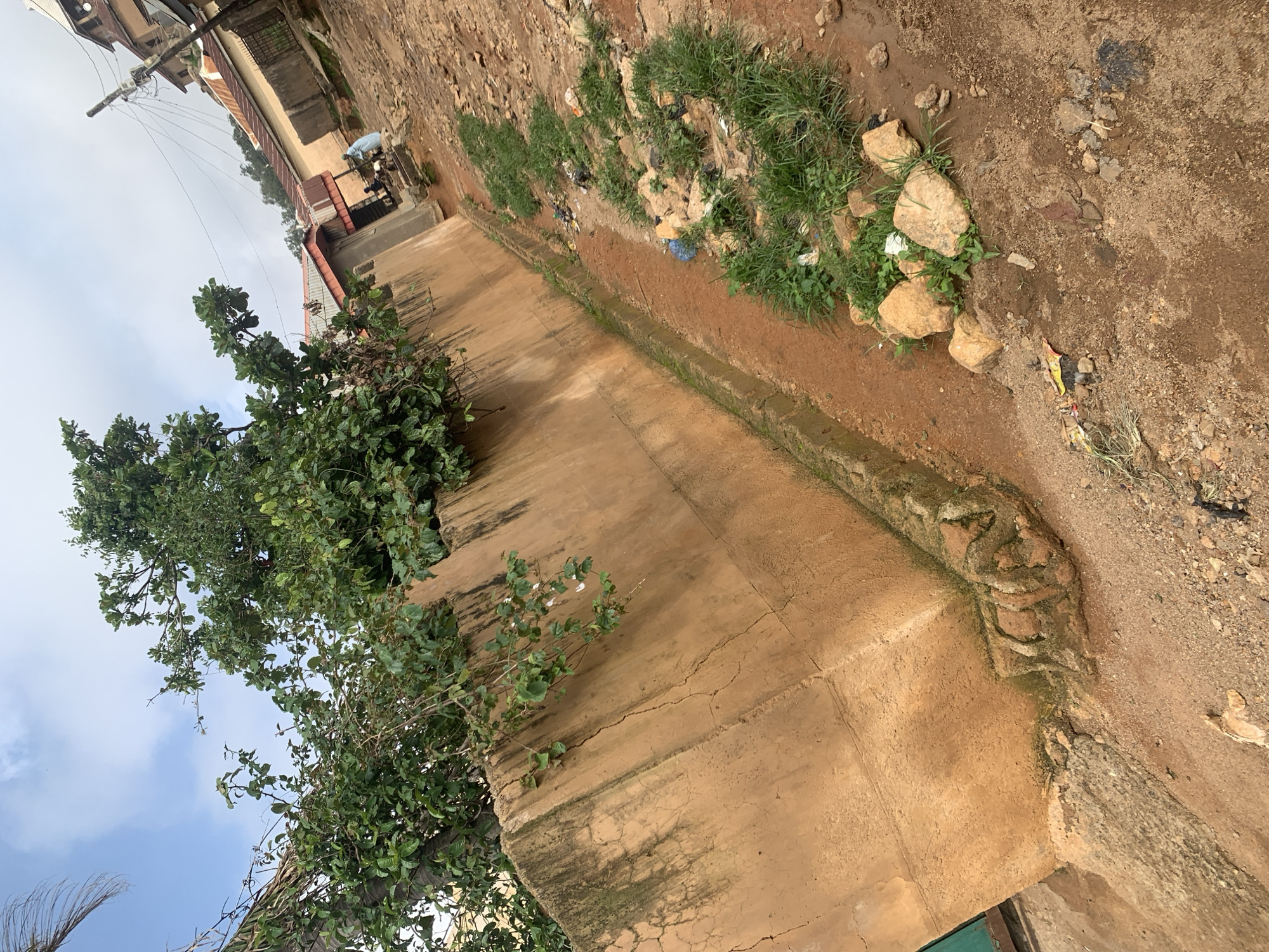 The image depicts a street scene in Jos, Nigeria. A weathered, ochre-colored concrete wall runs diagonally across the frame, partially obscured by lush green foliage from trees growing along its top. To the right of the wall, a dirt road is visible, with a shallow, overgrown drainage ditch running alongside the wall. Scattered across the road and in the ditch are rocks, patches of grass, and some litter, including plastic bags and wrappers. In the background, several buildings with corrugated metal roofs are visible, along with a utility pole and electrical wires. A person wearing a light blue garment is standing near a building in the middle ground. The sky above is partly cloudy.