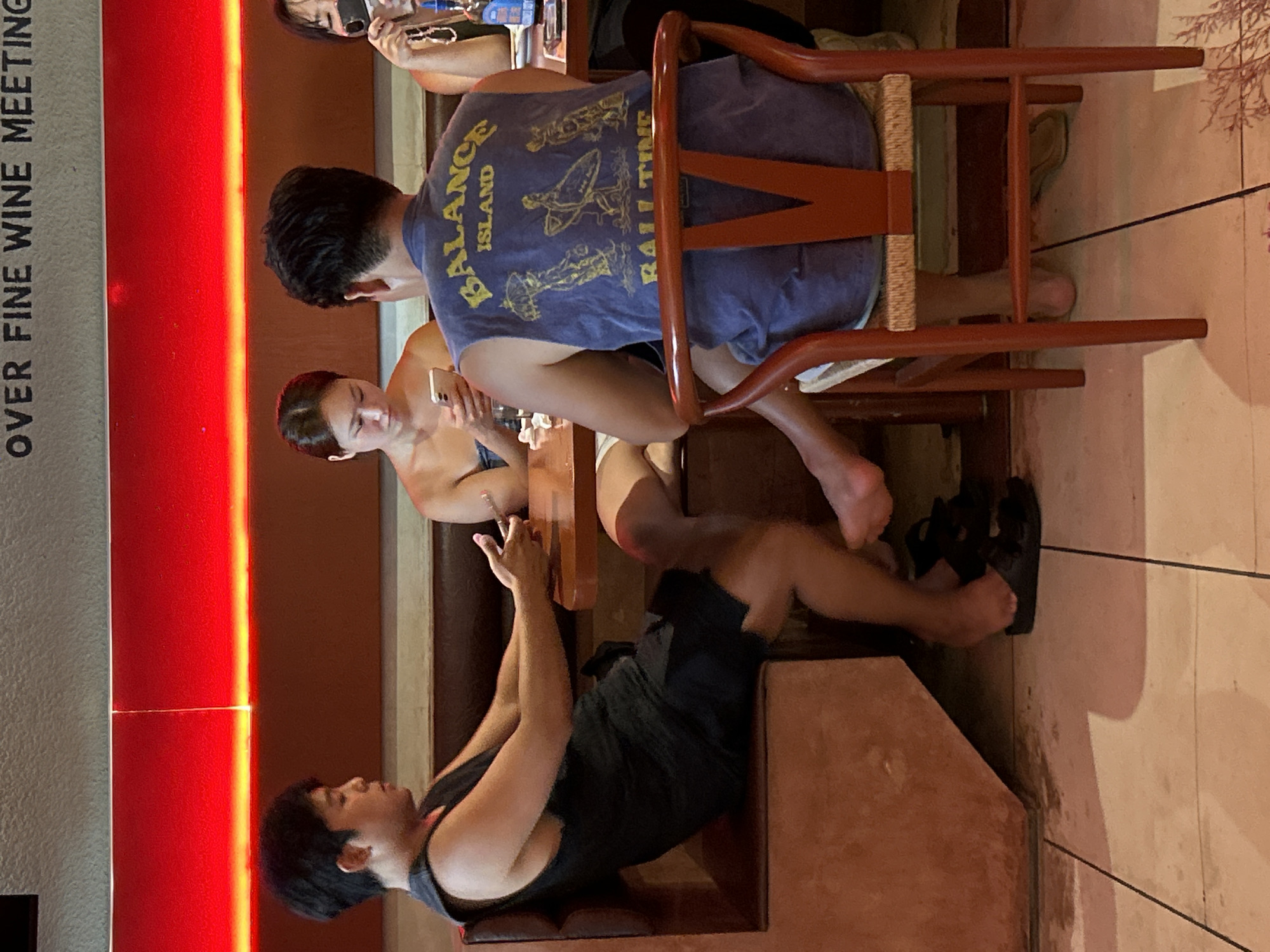 A group of people are gathered at a Pererenan, Indonesia venue that advertises "OVER FINE WINE MEETING." A man sitting on a chair wears a blue t-shirt with "BALANCE ISLAND" and "RAIL TIME" printed on the back. A woman seated next to him is looking at her phone. Another man is seated at the edge of the frame, facing them.