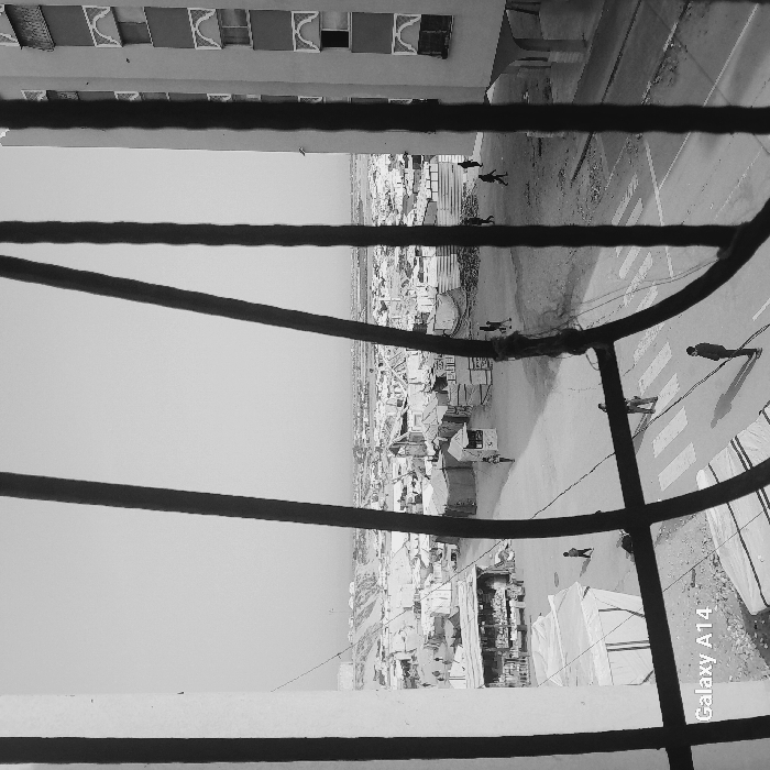 A black and white photo depicts an urban scene through a barred window. The view includes a street with pedestrian crossings and several figures walking. Beyond the street, a sprawling cityscape extends into the distance under a bright sky. The window bars frame the scene, offering a textured foreground. The building the window is attached to is partially visible in the upper left corner. The image is stamped with "Galaxy A14".