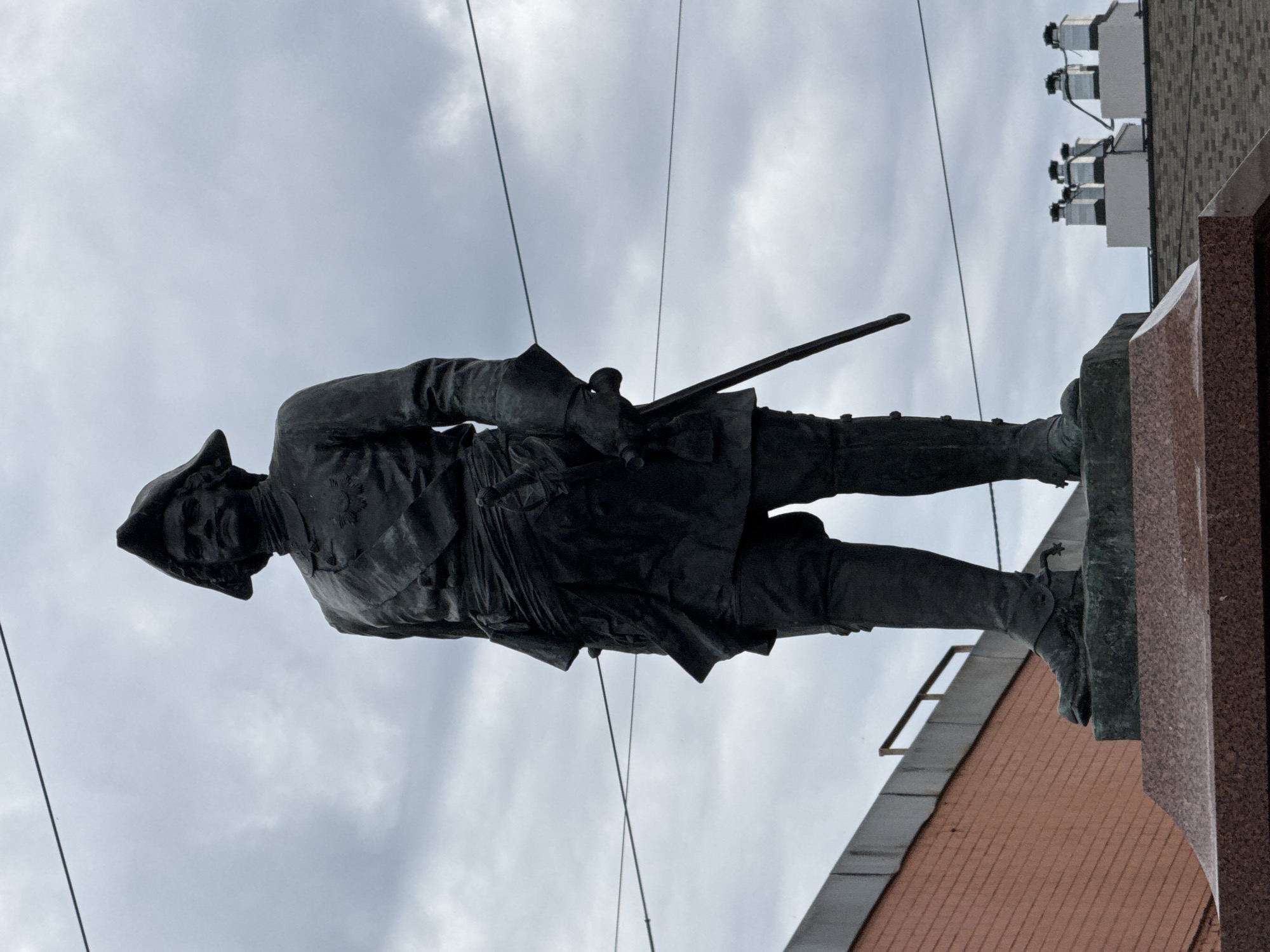 The image depicts a full-body bronze statue of Peter the Great, a historical figure of Russia, positioned on a pedestal. The statue is oriented facing forward, with the subject's head turned slightly to the left. Peter the Great is depicted wearing a tricorn hat, a uniform with a sash and a sword at his side. His right hand is resting on the pommel of his sword. The statue is situated outdoors against a cloudy sky. In the background, a red brick building is visible on the left, and on the right, a series of white chimneys with black caps stand atop a dark-tiled roof. The statue is located in the city of Saint Petersburg, Russia, specifically at the Bronze Horseman monument.