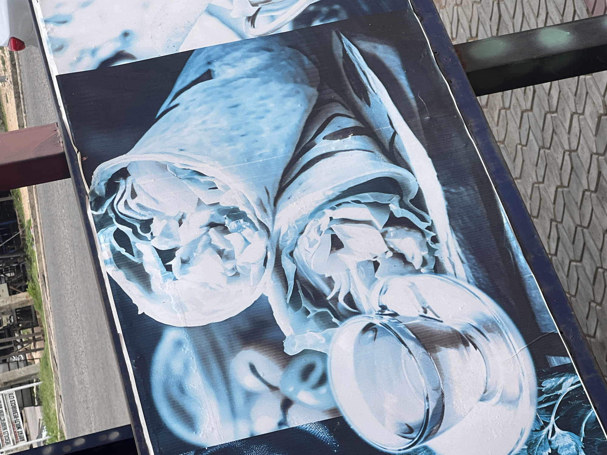 An outdoor advertisement board in Jalingo, Nigeria, displays a close-up, monochromatic blue image of two wraps and a cup of coffee on a saucer. The wraps appear to be filled with shredded lettuce and possibly chicken or other filling. The cup is clear glass, with a reflection of the surroundings visible on its surface. The advertisement is mounted on a dark blue metal stand. In the background, a paved street is visible, with some grass and a white sign on a pole. A white car is parked in the distance. The ground is paved with interlocking bricks.