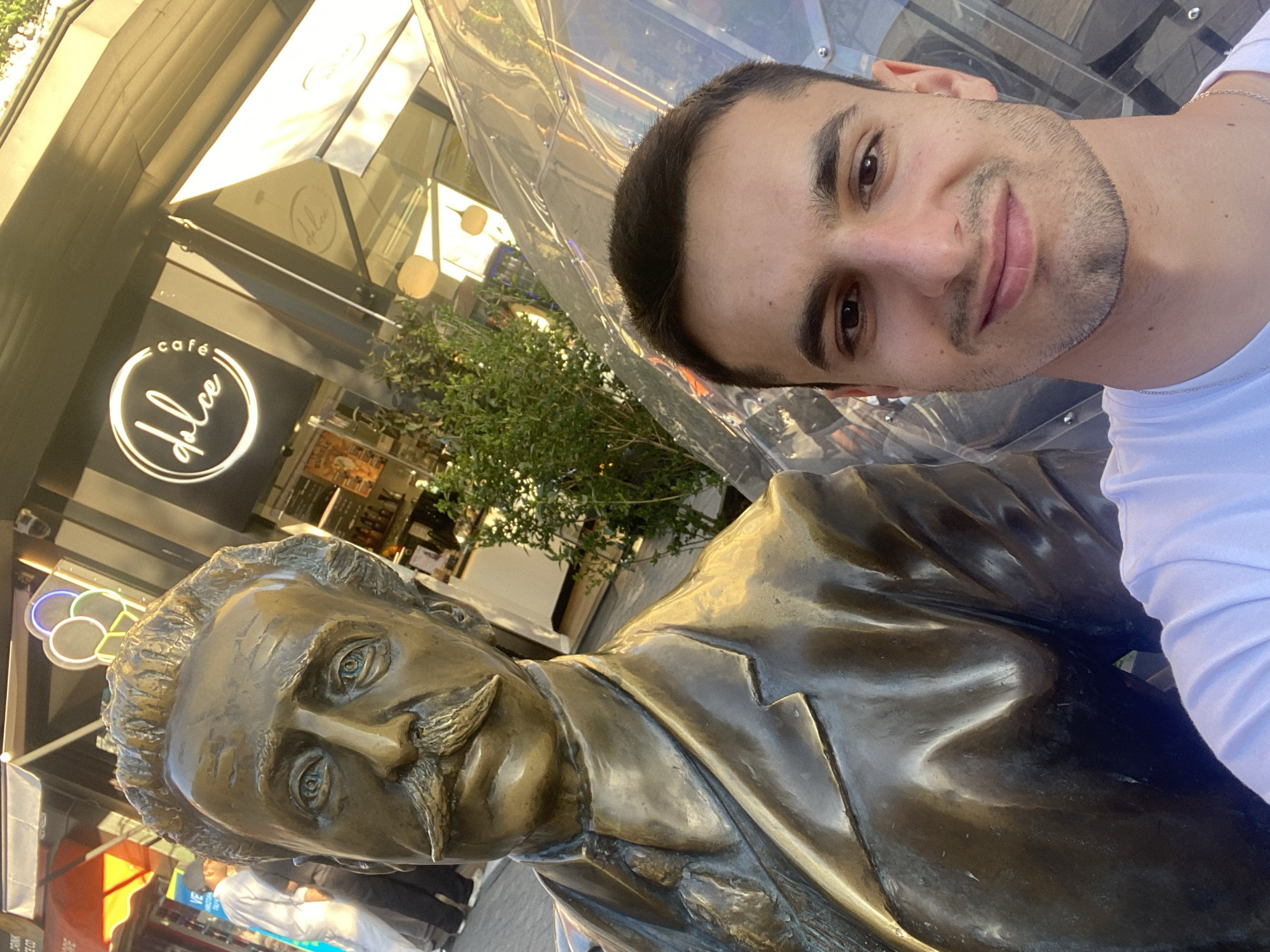 A selfie taken at an outdoor cafe in Varna, Bulgaria, features a young man with short dark hair, brown eyes, and a slight beard wearing a white t-shirt, positioned next to a bronze statue of a historical figure with a mustache. The statue is wearing a suit jacket. The cafe's sign reads "cafe dolce" and there is an outdoor seating area with clear plastic enclosures. The environment appears to be sunny and urban.