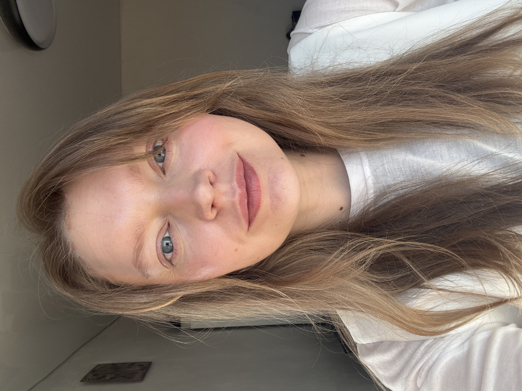 A close-up portrait of a woman with light brown hair and blue eyes. She is wearing a white top. There's no evidence indicating the location is Izhevsk, Russia. The background is a plain wall.