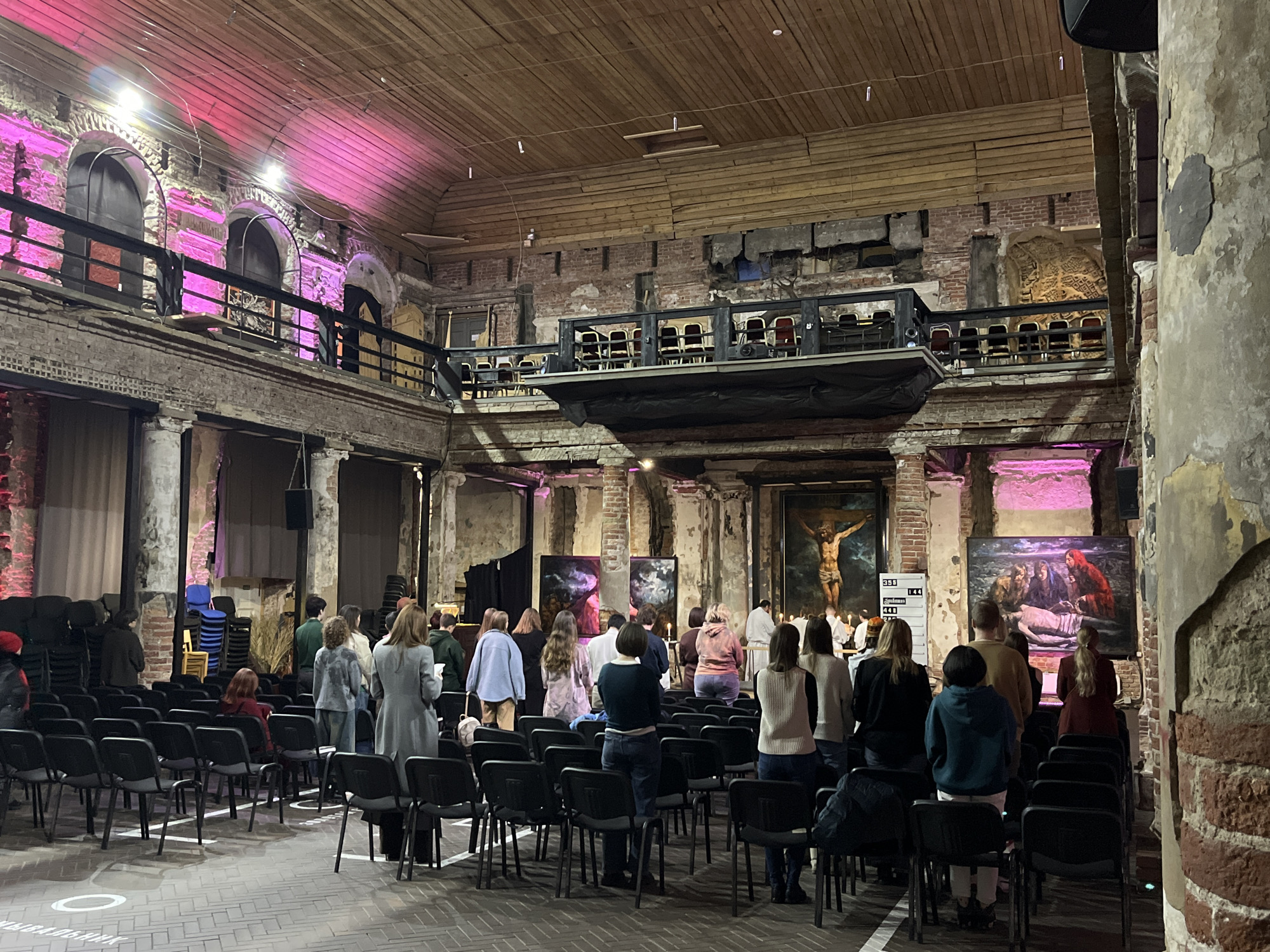 The scene depicts an indoor gathering within the Annakirche (St. Anna's Church) in Saint Petersburg, Russia. The large, multi-level hall features distressed red brick walls, peeling plaster, and large, worn columns, contrasting with a high, pitched wooden plank ceiling. A cantilevered balcony with dark metal railings and chairs extends into the space from the right, while a higher gallery with arched openings and similar railings is visible on the left, illuminated by pink uplighting. The main floor is paved with a light-colored brick pattern, marked with white social distancing circles and lines.
Approximately 30-40 individuals are present, mostly standing in the foreground and middle ground, facing the front of the hall. They are diverse in age and casually dressed; some hold small pamphlets. At the front, several large paintings are displayed: a prominent depiction of the Crucifixion of Christ centrally, flanked by a painting of a Lamentation scene to its right and two abstract artworks to its left. Lit candles are visible near the base of the central artworks. A small group of people, some appearing to be officiants in white robes, stand at the very front near the paintings. A white display board with black numbers (358, 144, 448) is positioned to the left of the Crucifixion painting. Large dark speakers are mounted high on the walls. Black stackable chairs are arranged in rows on the main floor, with additional stacked chairs visible in the background.