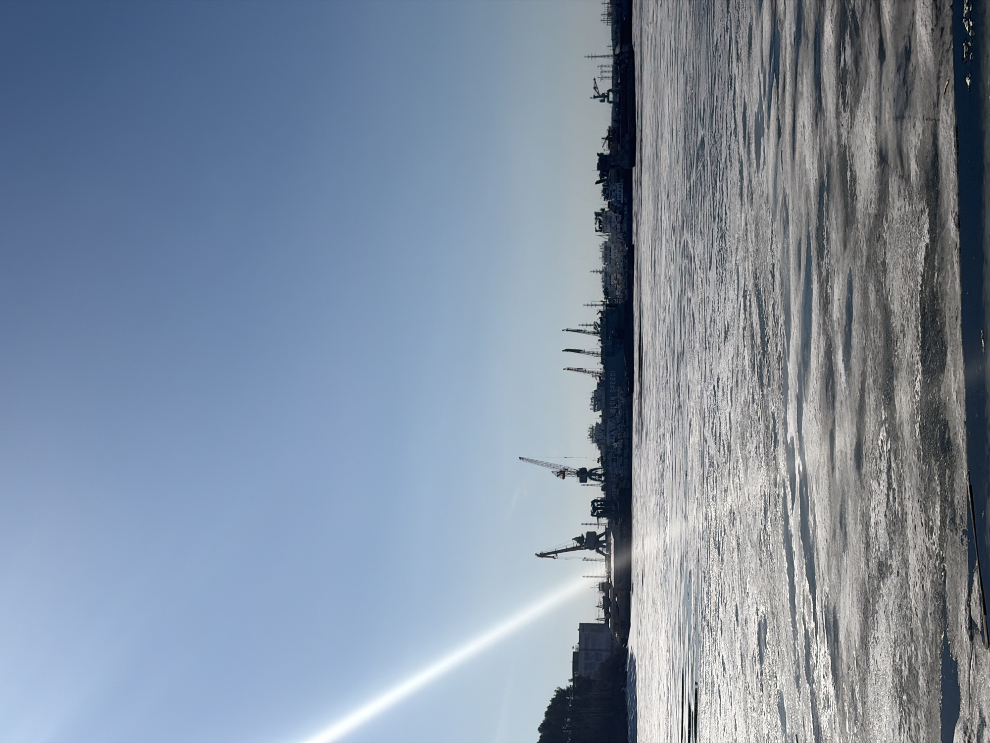 This panoramic image, correctly oriented, depicts a vast, frozen body of water under a clear blue sky. The scene is dominated by a large expanse of ice in the foreground and midground, characterized by distinct ridges and textures, suggesting a river or lake frozen solid during winter.
Along the distant horizon, a dense line of industrial structures is silhouetted against the bright sky. Numerous cranes of varying sizes and shapes are prominent, indicative of a port, shipyard, or an active industrial waterfront. Several large vessels or structures are also visible among the cranes.
The sky is a bright, uniform blue, with a subtle gradient from a lighter hue near the horizon to a deeper blue overhead. A strong lens flare emanating from the lower-left corner (when viewed correctly) suggests the sun is low in the sky, either rising or setting, casting bright light across the scene. The overall lighting implies a crisp, clear day, likely cold given the frozen water.
No specific activity is taking place; the scene is static and serene, capturing a moment in a cold, industrial landscape. The location context of Chaikovsky, Russia, aligns well with the visual evidence of a frozen river (like the Kama River) and industrial development along its banks.
Faint white text, "SLIM SPEED", is subtly visible on one of the larger industrial structures or ships in the background, near the upper part of the industrial line.
