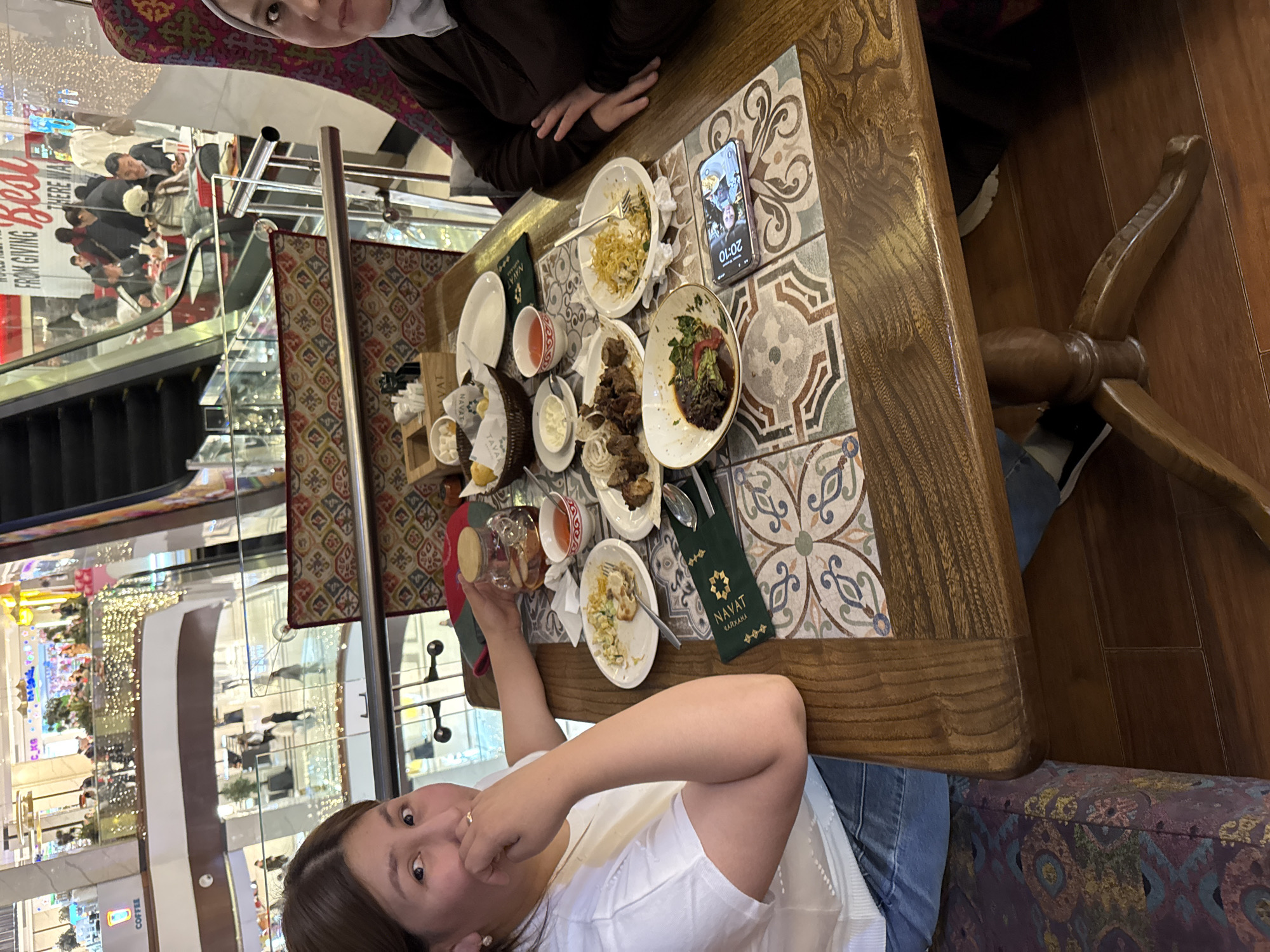 This image depicts an indoor scene at a restaurant within a bustling shopping mall. The perspective is slightly overhead, looking down at a wooden table where two women are dining.
In the immediate foreground, a woman in a white top and blue jeans is looking directly at the camera with a slight, perhaps playful, expression. She holds a transparent cup with a lid, seemingly drinking from it. Another woman, wearing a dark top and a light-colored hijab, is partially visible at the top of the frame, seated across the table.
The table itself is made of wood with an inset tiled section featuring ornate, colorful patterns. It is filled with several dishes and cutlery, indicating a recently concluded or ongoing meal. Visible items include plates with remnants of food such as noodles, meat, and salad, small bowls of sauces or condiments, a bread basket, and various cups. A green holder for cutlery, emblazoned with the text "NAVAT ЧАЙХАНА" (NAVAT Chaihana), is also on the table. A smartphone lies face up, displaying the time "20:10" on its lock screen, suggesting it's evening.
The background shows a brightly lit, multi-story shopping mall environment. Escalators, glass railings, and numerous shoppers are visible across different levels. Reflections off the glass surfaces distort some background elements. Partial text from store signs can be seen, including "FROM GIVING BE..." and "COFFEE". The overall setting suggests a vibrant commercial and dining experience, consistent with a modern shopping center in Bishkek City, Kyrgyzstan.