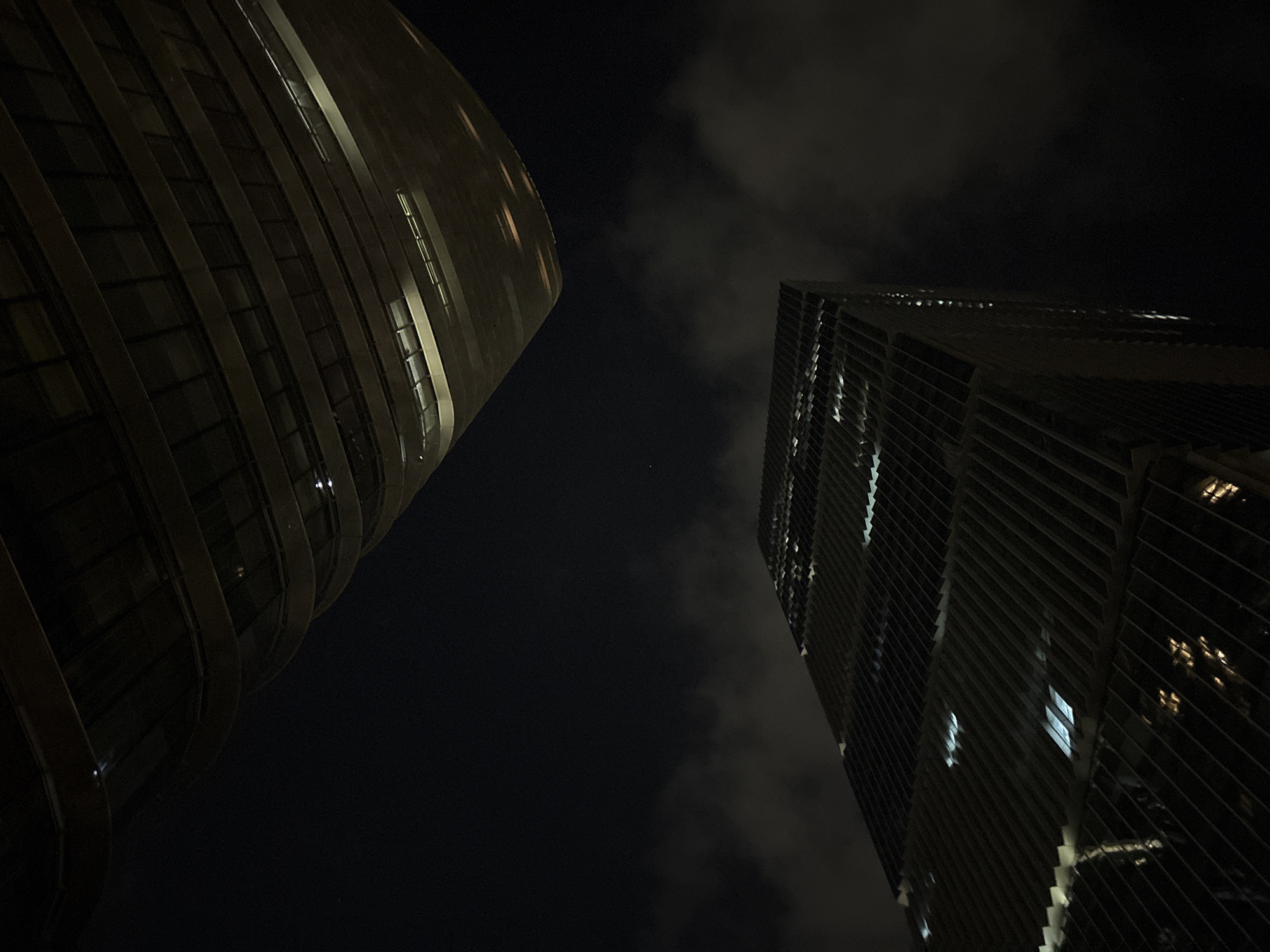 This media file shows a low-angle view of two modern skyscrapers at night. The building on the left has a curved, rounded facade with horizontal banding and illuminated windows. The building on the right is taller and rectangular, with a facade characterized by vertical and diagonal louvers or fins. The windows of this building are also lit, some appearing as bright white rectangles. The sky is dark and cloudy, with some of the clouds illuminated by the city lights. A small, bright speck, possibly a distant star or a very distant light, is visible in the sky between the two buildings. The overall impression is of an urban nighttime scene with dramatic architecture. The location context provided is Haizhu District, China. There is no visible text in the image.