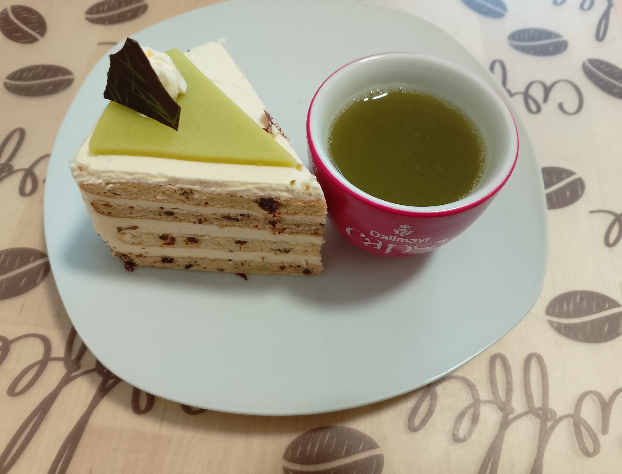 A slice of layered cake with green frosting and a chocolate garnish sits on a pale blue plate next to a red mug filled with green tea. The plate and mug are placed on a beige placemat with a coffee bean and script pattern. The scene is indoors, with no specific city or location identifiable.