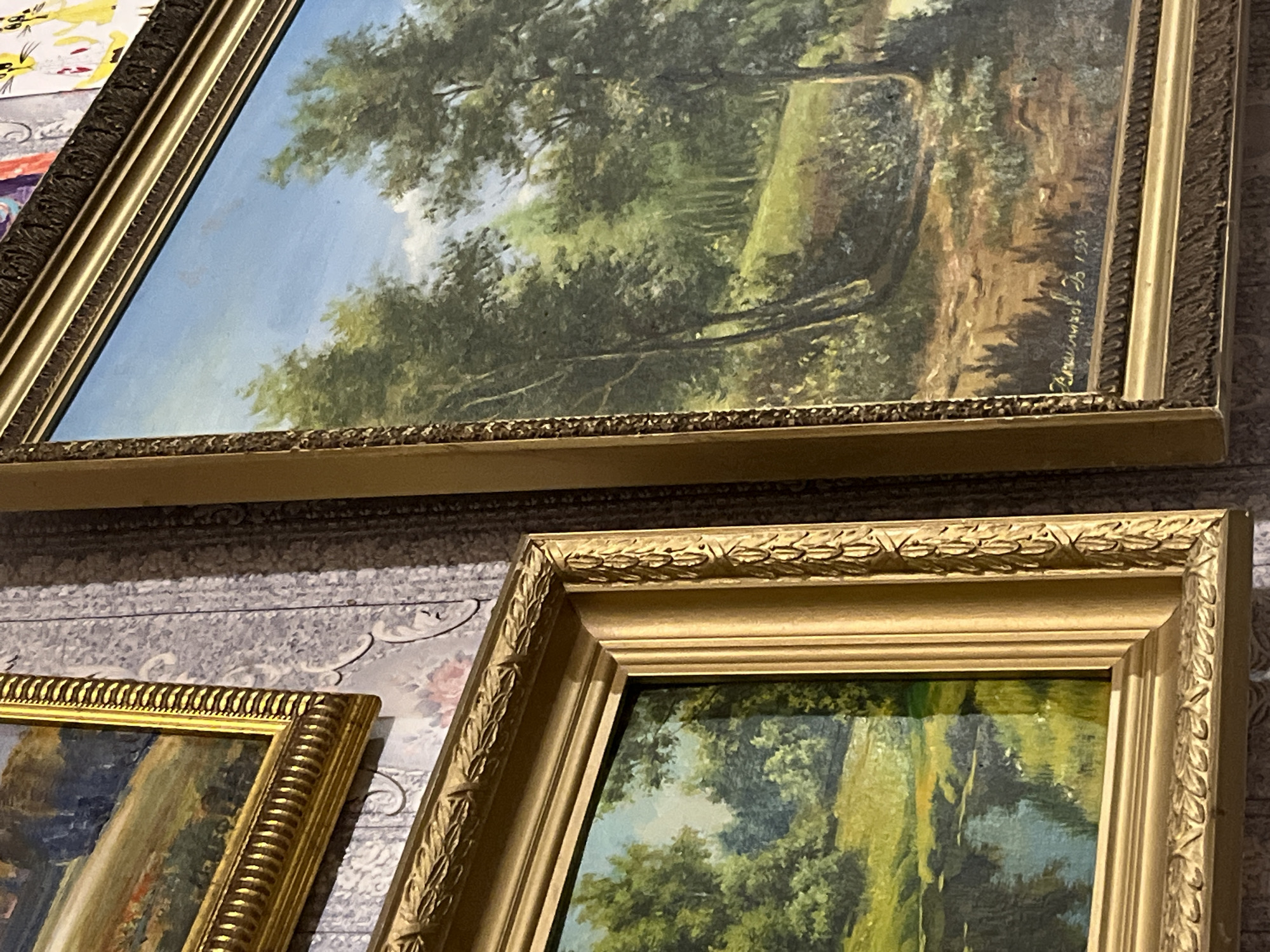 The image displays multiple framed landscape paintings, likely oil on canvas, positioned against a decorative, patterned wall. The central focus is on the uppermost painting, which depicts a tranquil, sunlit woodland scene. Dense green foliage of trees fills the canvas, with a bright path or clearing visible deeper within the woods. A clear blue sky with soft white clouds can be seen above the tree canopy.
This main painting is housed in an ornate, gilded frame, featuring intricate decorative patterns and beading along its edges. In the lower right corner of this artwork, a legible signature is visible, reading "Рудничнов З." (Rudnichnov Z.) followed by the date "1959," identifying the artist and the year of creation.
Below the prominent painting, sections of at least two other framed artworks are visible. These also feature highly decorative, gilded frames with distinct patterns; one partially seen frame on the lower left has a fluted or beaded design, while another on the lower right showcases a relief pattern resembling laurel leaves along its outer edge. These partial views suggest they are also landscape paintings consistent with the style of the main piece.
The paintings are displayed indoors, leaning against a backdrop of light-colored, possibly grey and white, patterned wallpaper or fabric. The scene suggests a display setting, such as a gallery, antique shop, or a private collection.