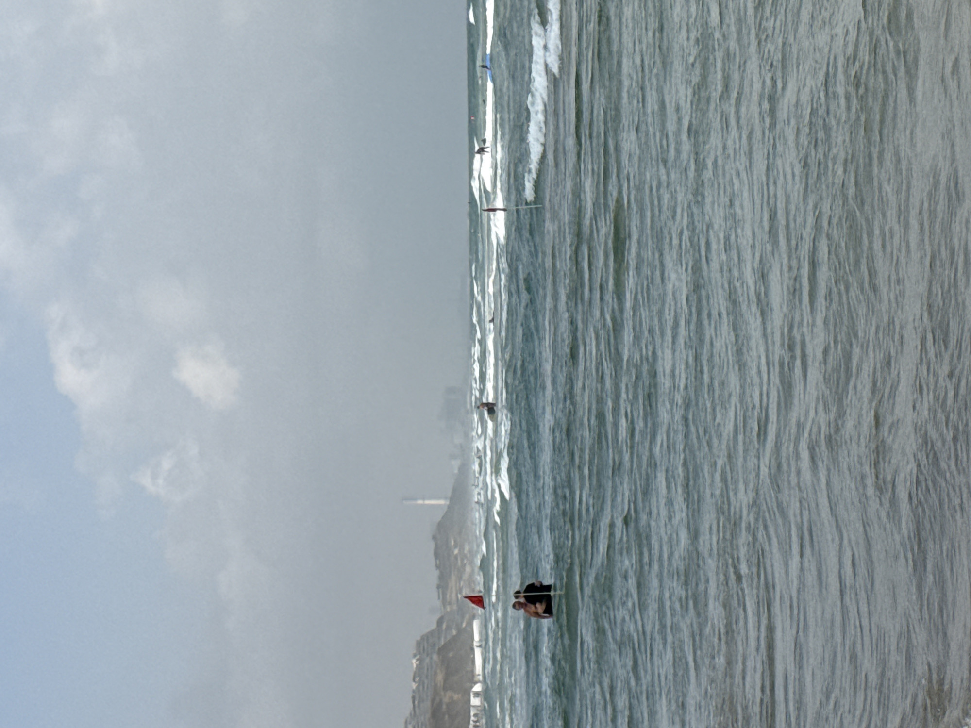 This image captures a portion of the coastline in Herzliya, Israel, under a cloudy sky. The foreground is dominated by choppy ocean waves, with a faint shoreline visible in the distance.
In the middle ground, two individuals are standing in the water, partially submerged, with a red flag on a pole planted between them. Further out in the water, several other people can be seen, some appearing to be surfing or paddling on boards. A tall, slender structure resembling a radio tower or lighthouse is visible on the distant hills, along with buildings and greenery, indicating the presence of a developed area. The overall impression is a moderately busy day at the beach, with people engaged in water activities despite the overcast weather.