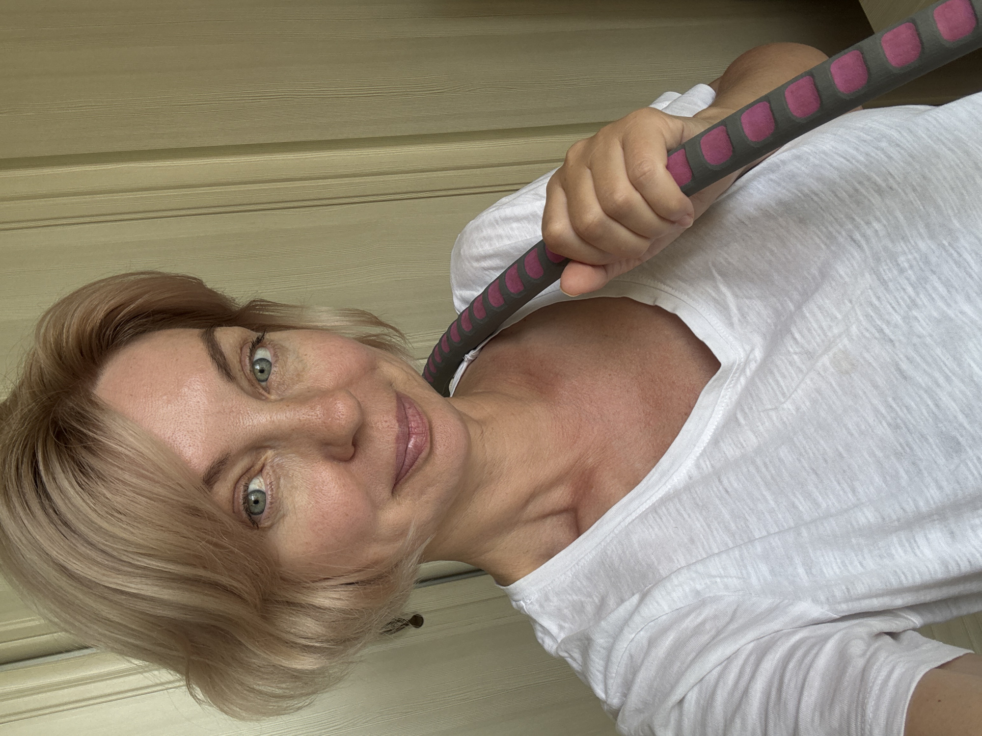 A medium shot captures a woman with blonde, wavy hair and light blue eyes looking directly at the camera. She is wearing a plain white t-shirt. Her right arm is bent, and her hand is holding a portion of a weighted hula hoop, which is gray with pink circular pads. The background consists of a pale yellow, vertically paneled door or wall. The lighting suggests an indoor setting, likely during the daytime. There is no visual information to suggest a specific location within Sochi, Russia.