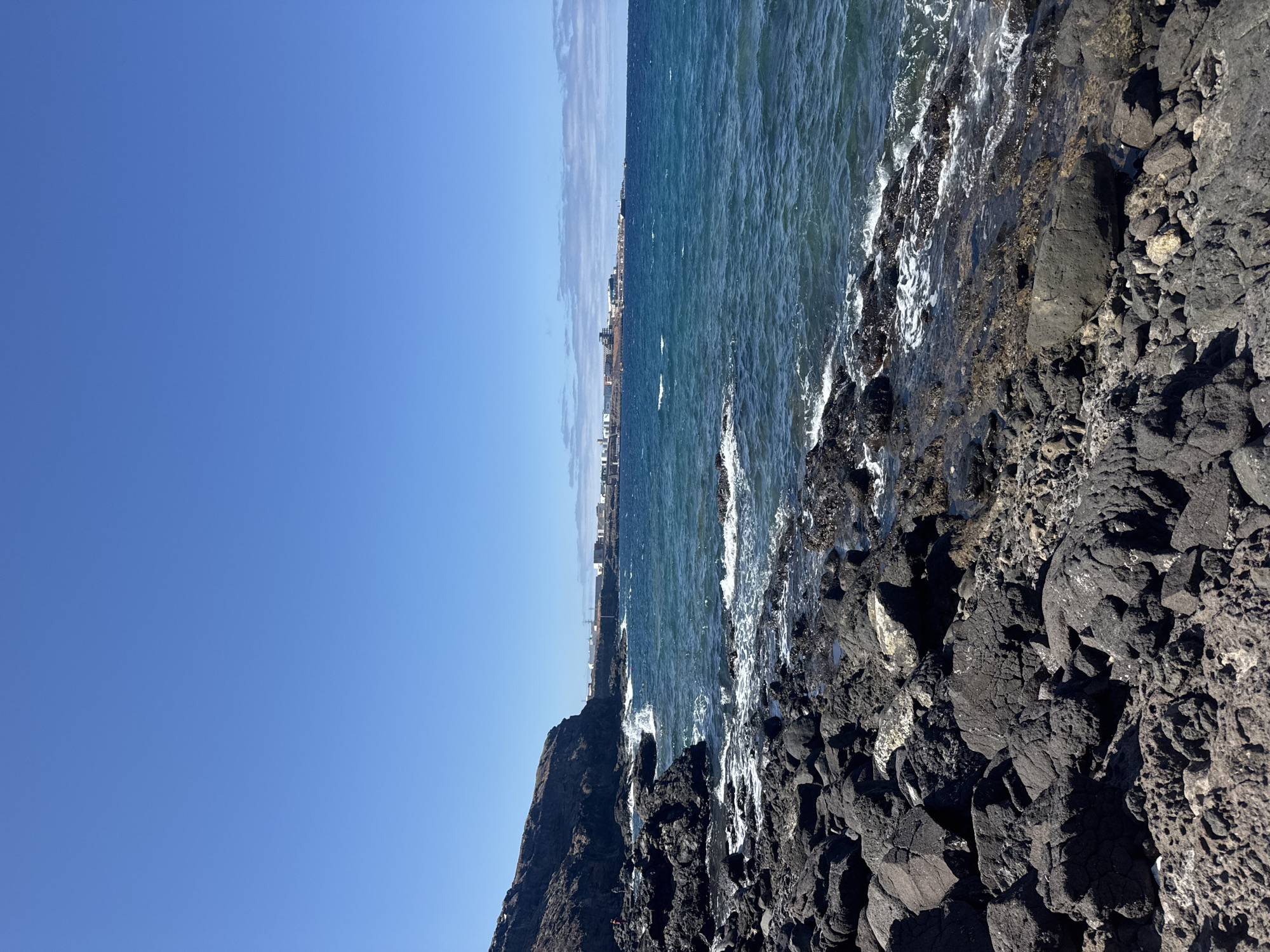 A vertical, eye-level, wide shot captures a rocky shoreline in Telde, Spain, on a clear, sunny day. The foreground is dominated by dark, rough volcanic rocks. Waves crash against the rocks, creating white foam. In the midground, the blue ocean stretches out, meeting the horizon. In the distance, a city skyline is visible along the coast, with buildings and structures under a bright blue sky. A cliff face is seen on the left side of the image.
