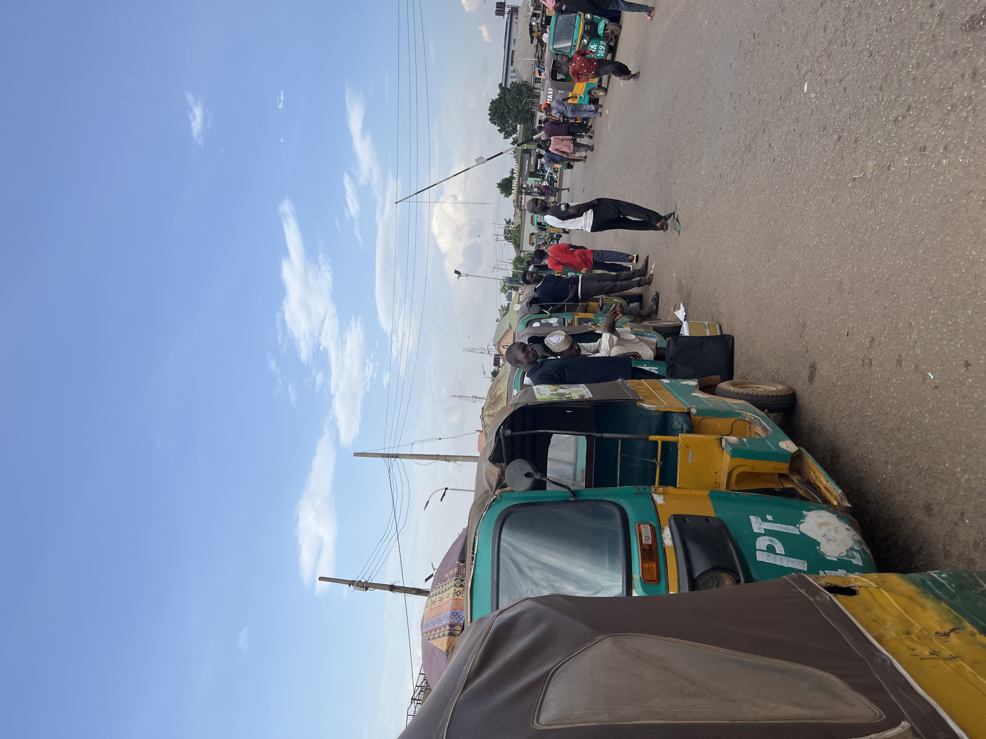 The image shows a row of parked auto-rickshaws (tuk-tuks) along a street in Gura Top, Nigeria. Several people are standing near the vehicles. The sky is blue with some clouds. Power lines are visible overhead.