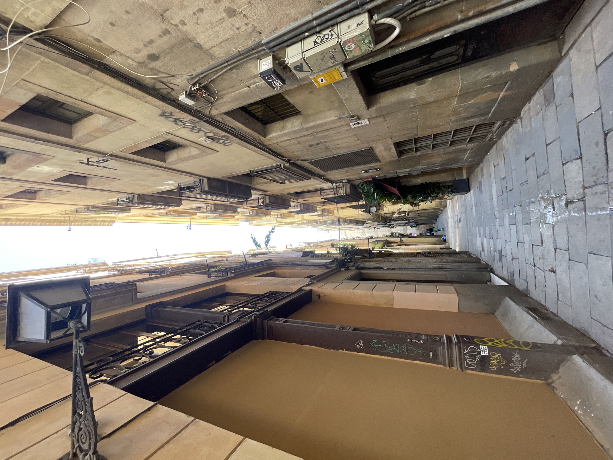 This image captures a very narrow urban alleyway or street in Barcelona, viewed looking dramatically upwards towards the sky. Tall, multi-story buildings flank both sides, creating a deep, canyon-like space with only a sliver of bright, clear sky visible at the very top, indicating daytime.
On the left, a building with light brown/ochre walls and darker architectural details features multiple windows, some adorned with decorative wrought-iron balconies. An ornate, black vintage-style lamppost extends from this building in the lower-left foreground. Patches of graffiti, including the word "ZODIAC," are visible on the lower parts of the building.
The right side of the alley presents another tall building. Its lower section features grey stone paving, suggesting the street surface. Above this, the building facade integrates various grates and utility installations. Overhanging elements between the buildings are dense with electrical conduits, wires, and junction boxes, many heavily covered in diverse graffiti. Visible text among the graffiti includes "SOHO STREET WEAR," "COPA," and "JOMAO." Further down the alley on the right, a "Restaurant" sign is visible, accompanied by some potted plants or foliage.
The architecture suggests an older, densely populated European city district. No people are visible.