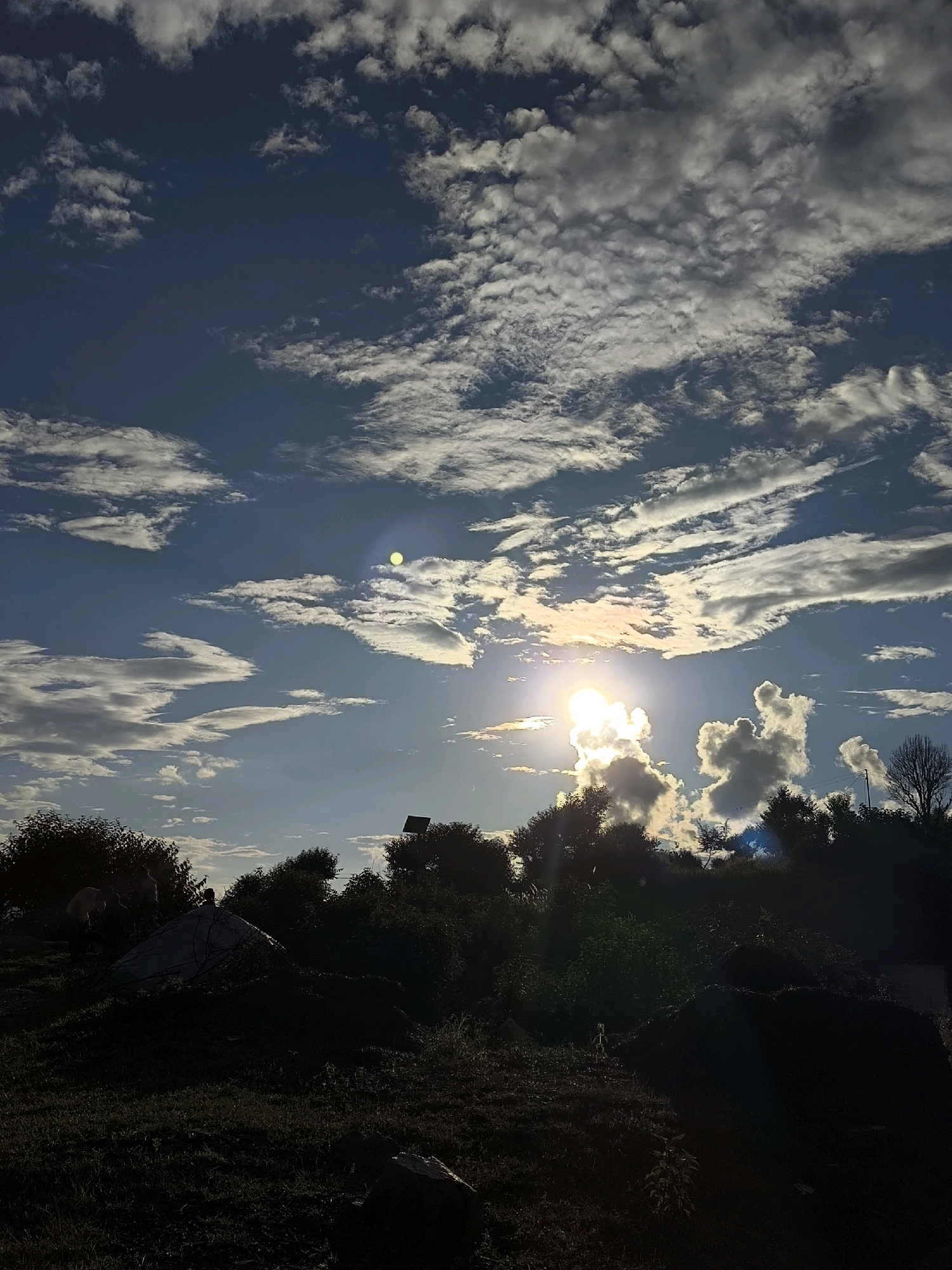 The image shows a silhouette of trees and hills against a bright sky. The sun is visible and is shining through the clouds. There is a lens flare in the upper right portion of the image. The foreground is dark and appears to be a grassy area with some rocks. The image is taken from a low angle, looking up at the sky.