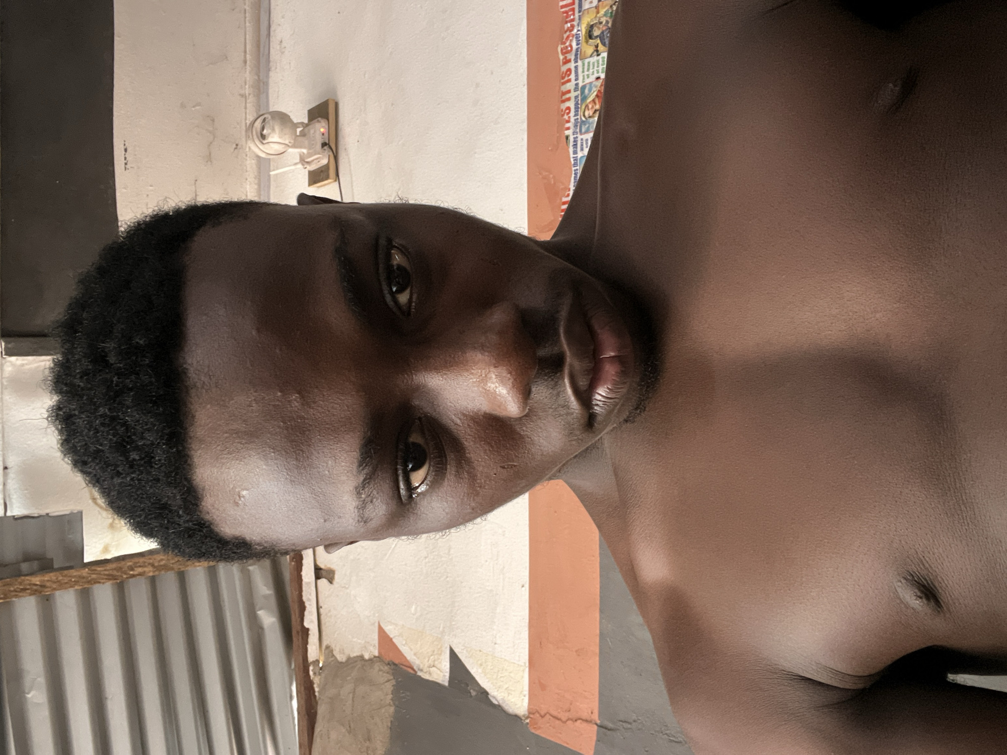 A shirtless young Black male is captured in a selfie-style photograph, his gaze directed intently at the camera. His hair is short and neatly styled. The background suggests an indoor setting with an unfinished ceiling made of corrugated metal and wooden beams. The wall behind him is textured and painted white, with distinct orange and gray stripes. A small, white electronic device, possibly a security camera or Wi-Fi router, is mounted on the wall. Posters with text and images are visible on the wall to the right of the subject. The lighting appears to be natural, casting a soft glow on his skin. The overall scene suggests a casual, possibly domestic, environment.