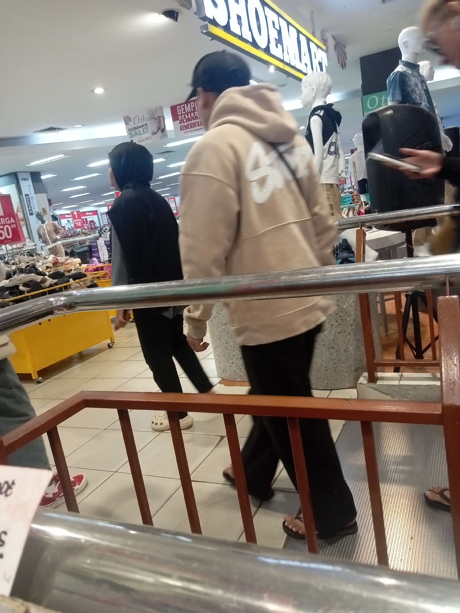 The image depicts an indoor environment, likely a shopping mall, with a focus on a retail store selling footwear. A sign above the store reads "SHOEMART". In the foreground, two individuals are partially visible, walking on a tiled floor. One person is wearing a light-colored hooded sweatshirt and black pants, while the other is wearing a black hijab and black pants. To the left, a yellow display rack holds shoes, and a poster with "HARGA 50.000" is visible. Mannequins are present in the background, displaying clothing. The lighting suggests an artificial indoor setting. The overall scene is consistent with a commercial retail space in Batam, Indonesia.