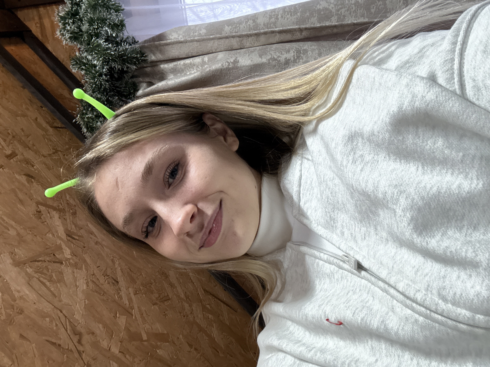A close-up selfie depicts a young, fair-skinned person with long, blonde hair partially visible, wearing a white zip-up hooded sweatshirt over a white turtleneck. Two green, antennae-like hair clips are visible in the person's hair. The background includes a wooden panel wall, a curtained window, and a decorative green garland. The location within the city of Прикамский, Russia cannot be determined from the provided image.