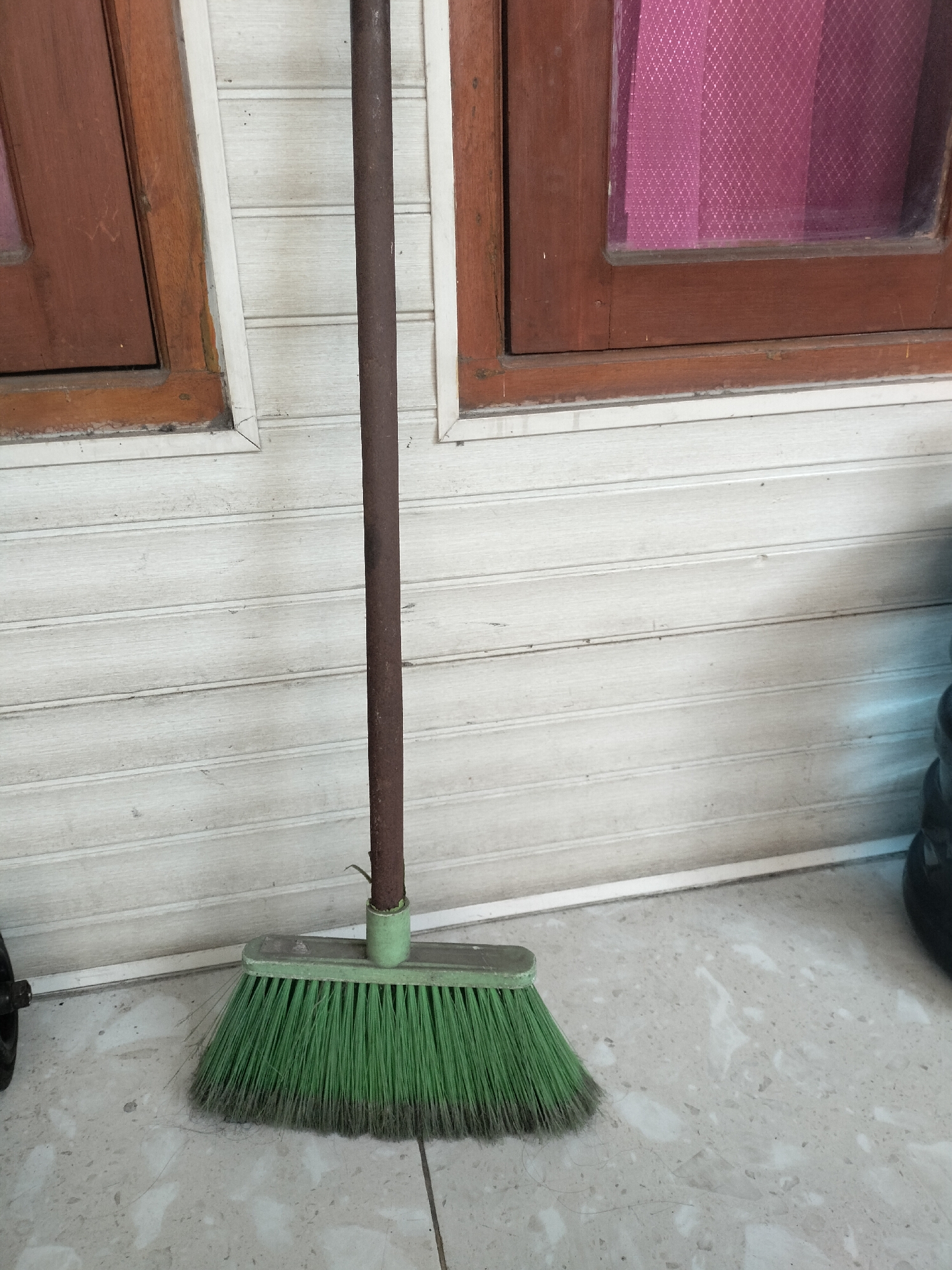 A broom with a green brush and a dark brown handle rests against a light-colored horizontal paneled wall. The broom is positioned in the foreground, with its brush spread out on a tiled floor. Two wooden-framed windows are visible above the broom, with the left window appearing to have a dark reddish-brown frame and the right window also having a similar frame and a pink, patterned glass. The floor is tiled with a speckled pattern in shades of grey and white. The environment suggests an outdoor or semi-outdoor setting, possibly a porch or patio area in Sidoarjo, Indonesia, given the architectural style and tiling.