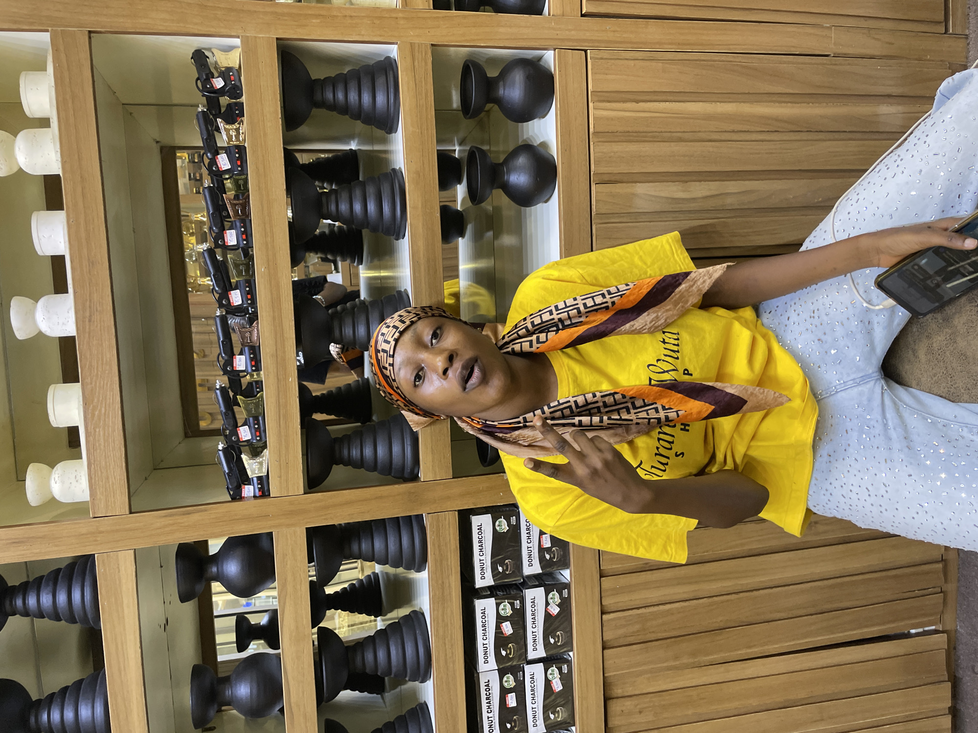 The image shows a young woman in a yellow t-shirt and light blue jeans, sitting in front of a display of products. She is wearing a headscarf with a patterned design. The shelves behind her are stocked with various items, including what appear to be decorative urns or vases in black and white, and rows of small boxes with text that includes "DONUT CHARCOAL." The woman is holding a smartphone in her right hand and is making a peace sign with her left hand. She has an open-mouthed expression, possibly speaking or laughing. The setting appears to be an indoor retail space, with wooden shelving and warm lighting, suggesting it could be a shop. The text visible on the boxes and on the woman's t-shirt ("Turan Wuta") provides some context about the products and potentially the brand. The overall mood seems casual and friendly. The location context provided, Unguwar Sarki, Nigeria, further narrows down the setting.