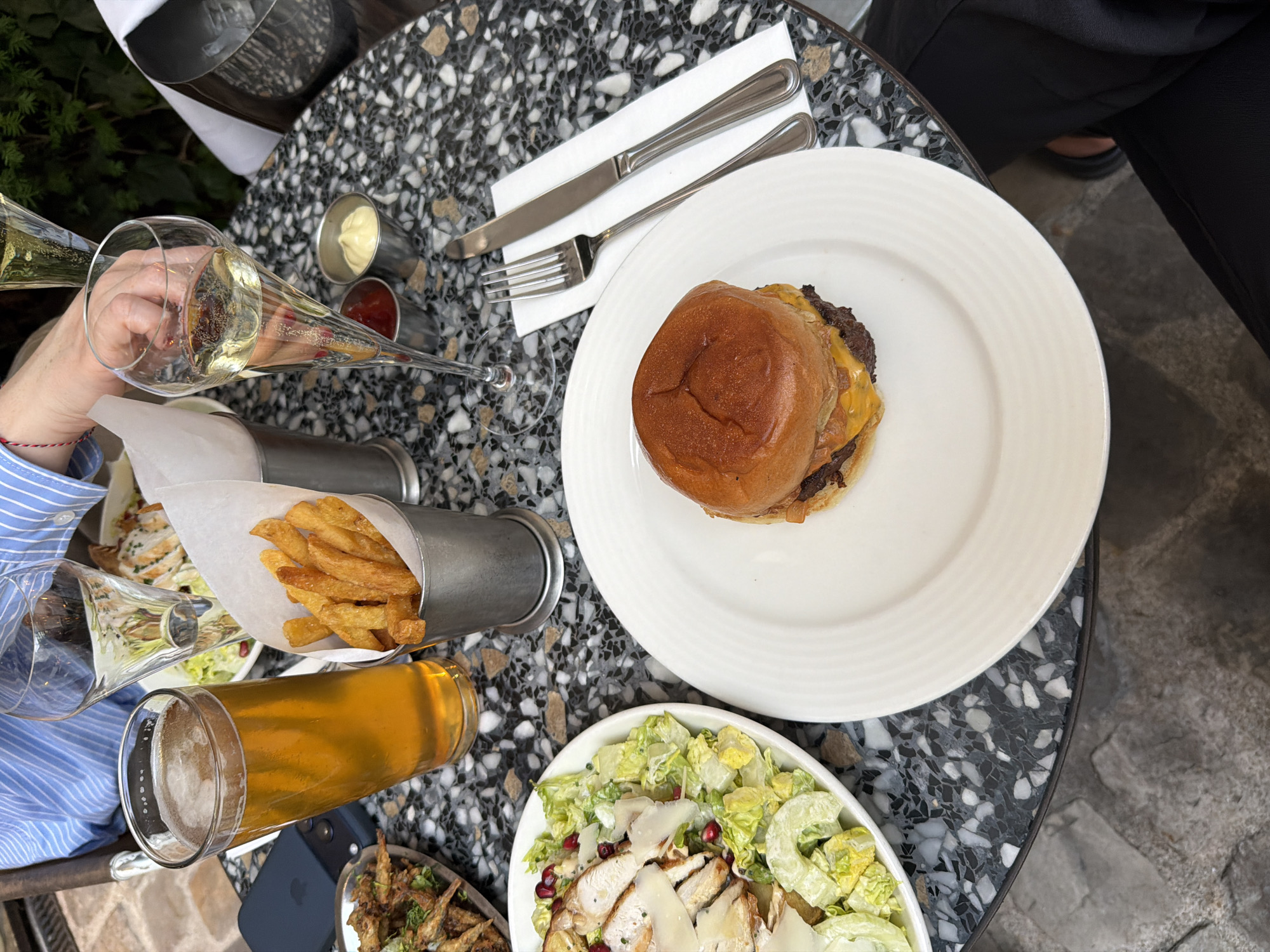 A table with a variety of food items is seen from an overhead perspective. The table has a speckled black and white pattern. The food items include a burger on a white plate, a salad with chicken, fries in a metal container, and small fried fish. There is also a glass of beer and champagne in the scene. The setting appears to be a restaurant or cafe, based on the presentation of the food and the overall ambiance. The location of the city of Paris, France, cannot be verified based on the provided image content.