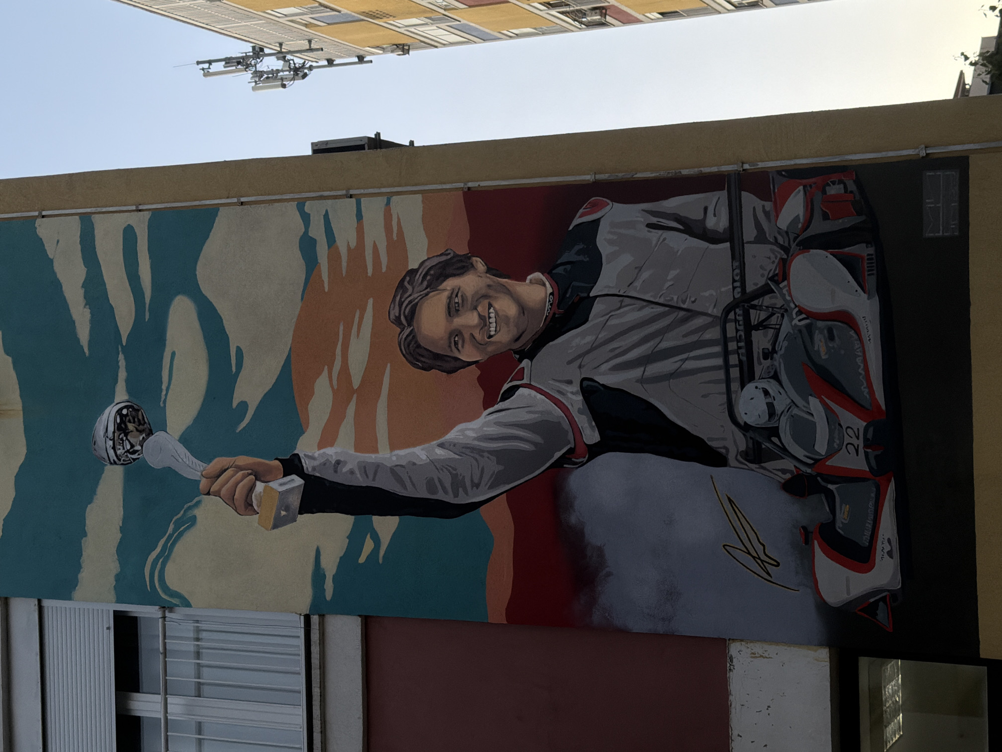 The image shows a mural on the side of a building. The mural depicts a man in racing attire holding a trophy. The background features abstract shapes and colors. A building with louvred windows is visible below the mural. In the upper portion of the image, parts of another building and an antenna-like structure are visible. The mural is of MotoGP rider Miguel Oliveira, located in Podgorica, Montenegro.