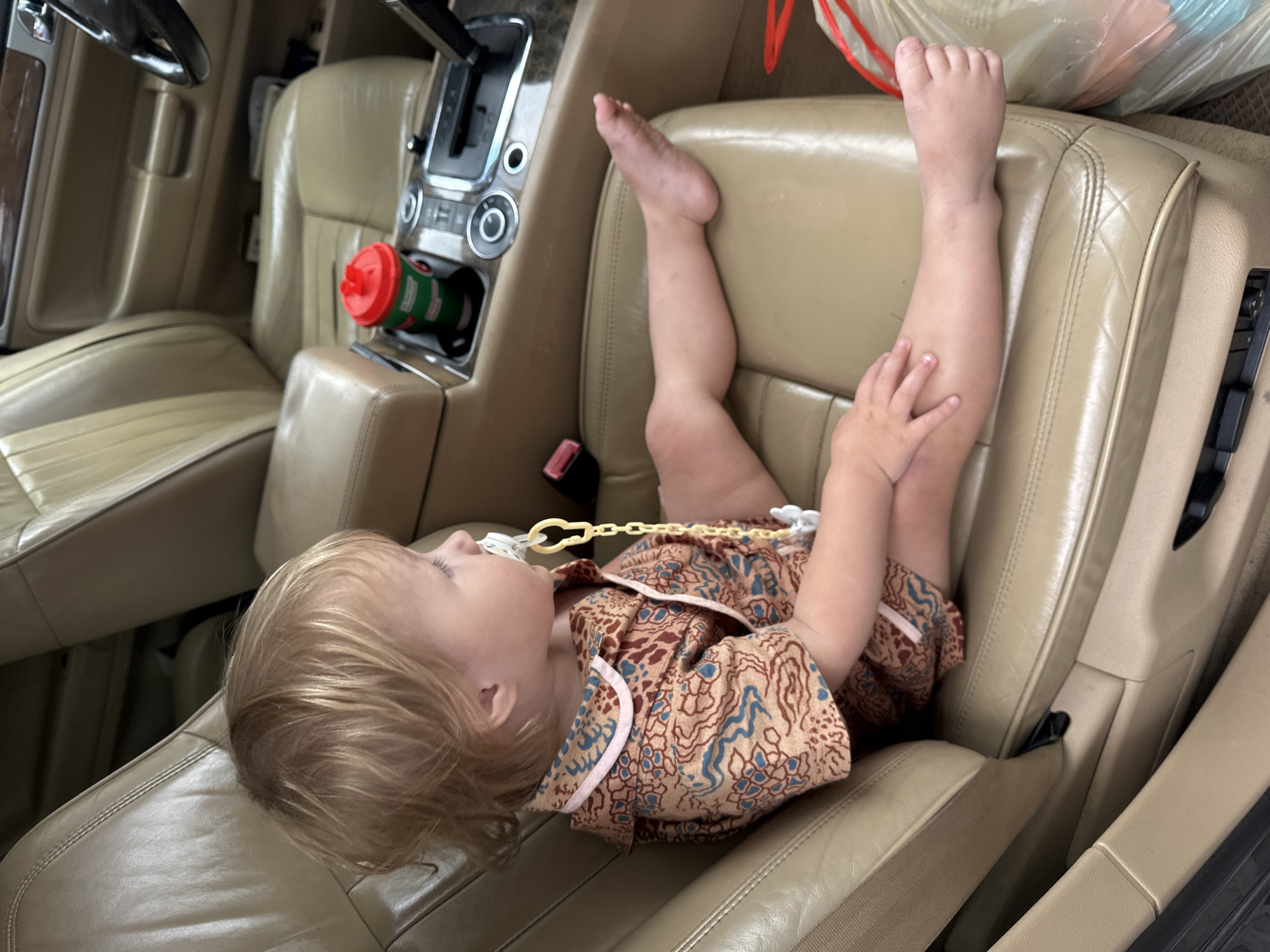 A young child with blonde hair is sitting in the passenger seat of a car, facing right. The child is wearing a light brown pajama set with a dark red and blue pattern, and a yellow pacifier clip is attached to their clothing. Their legs are extended across the seat, and their feet are bare. The car's interior is visible, with tan leather seats and a center console with a cup holder containing a green and red cup. A steering wheel is partially visible on the right side of the image. The background shows the dashboard and the driver's seat area. The scene appears to be an indoor setting, possibly a parking garage, given the lighting and the vehicle's environment. This image was taken in Nizhny Novgorod, Russia.