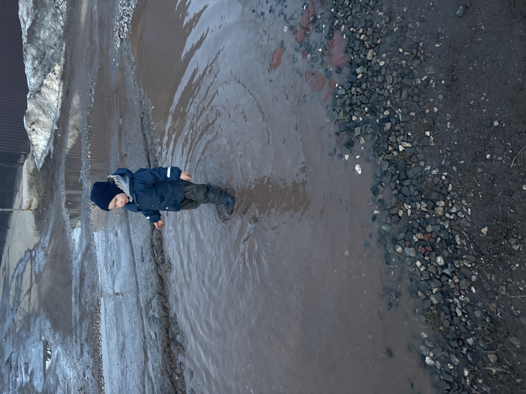 A child, wearing a dark blue jacket, dark pants, and dark boots, stands in a large puddle on a dirt and gravel road. The child is facing to the right of the frame. The puddle has concentric ripples emanating from the child. To the right of the puddle is a gravelly dirt area. To the left of the puddle is a dirt road. In the far left of the frame, there is a corrugated metal building or fence. The location is Ферма, Russia.