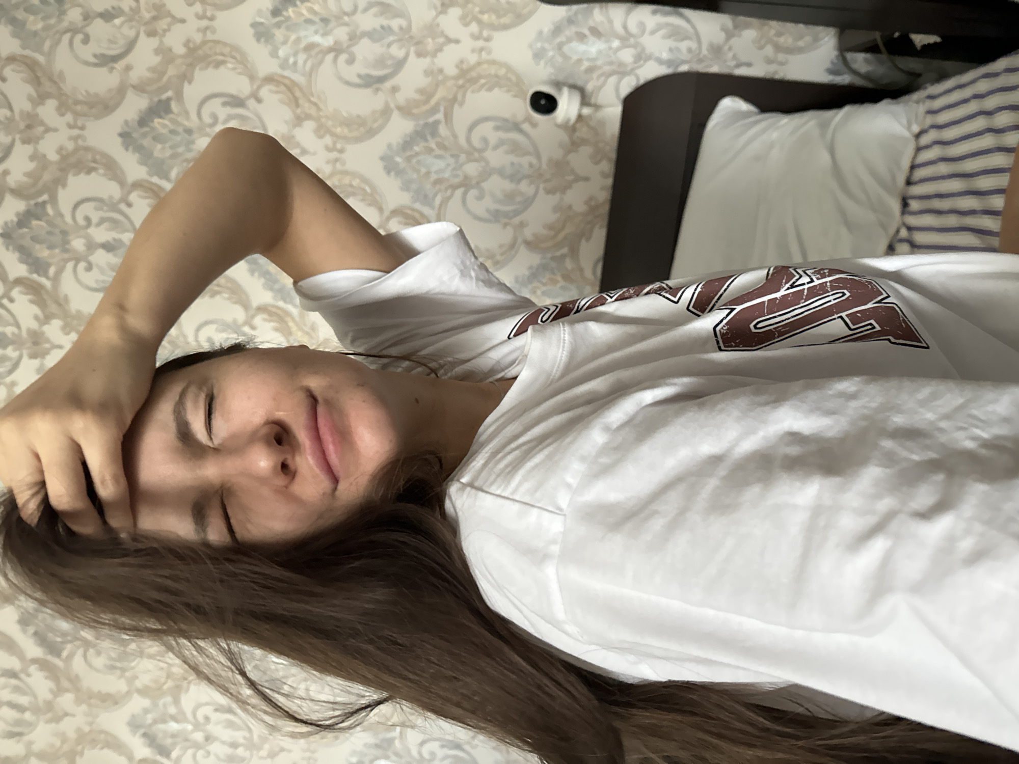 A woman is lying on a bed in a room with wallpaper featuring a beige and gray floral pattern. Her head rests on her right hand, with her eyes closed. She has long brown hair and is wearing a white T-shirt with a maroon design. The bed is made with a white pillow and a striped bedsheet. A white security camera is mounted on the wall behind the bed. A dark brown headboard is visible. The location in the city of Unknown, Unknown, is not specified.