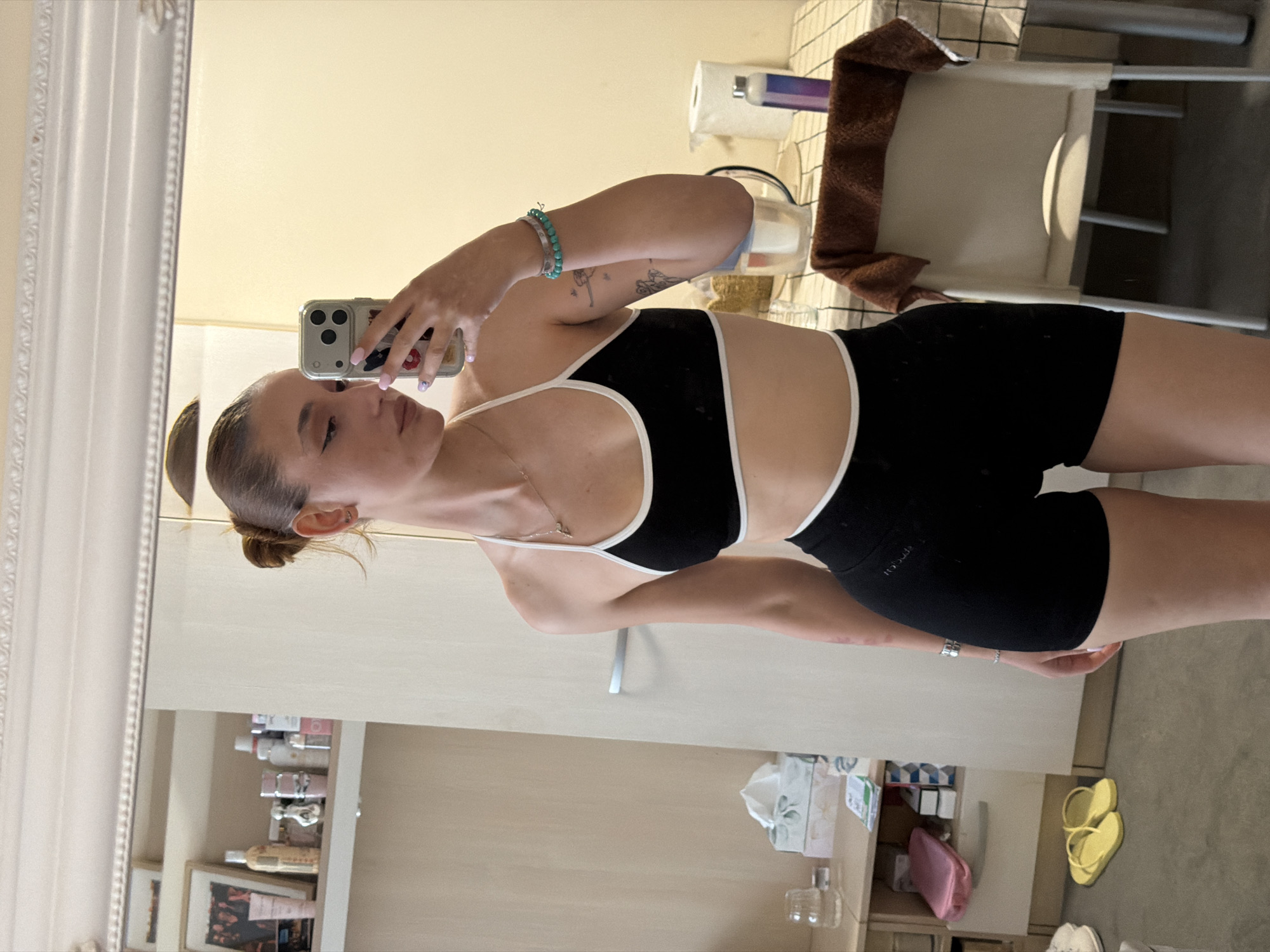 A woman is taking a mirror selfie in a bathroom. She has her hair in a bun and is wearing a black sports bra with white trim and black shorts. She is holding a phone with a white case. Behind her is a toilet, a sink, and a shelf of toiletries. There is no information available to determine if the location is Sofia, Bulgaria.