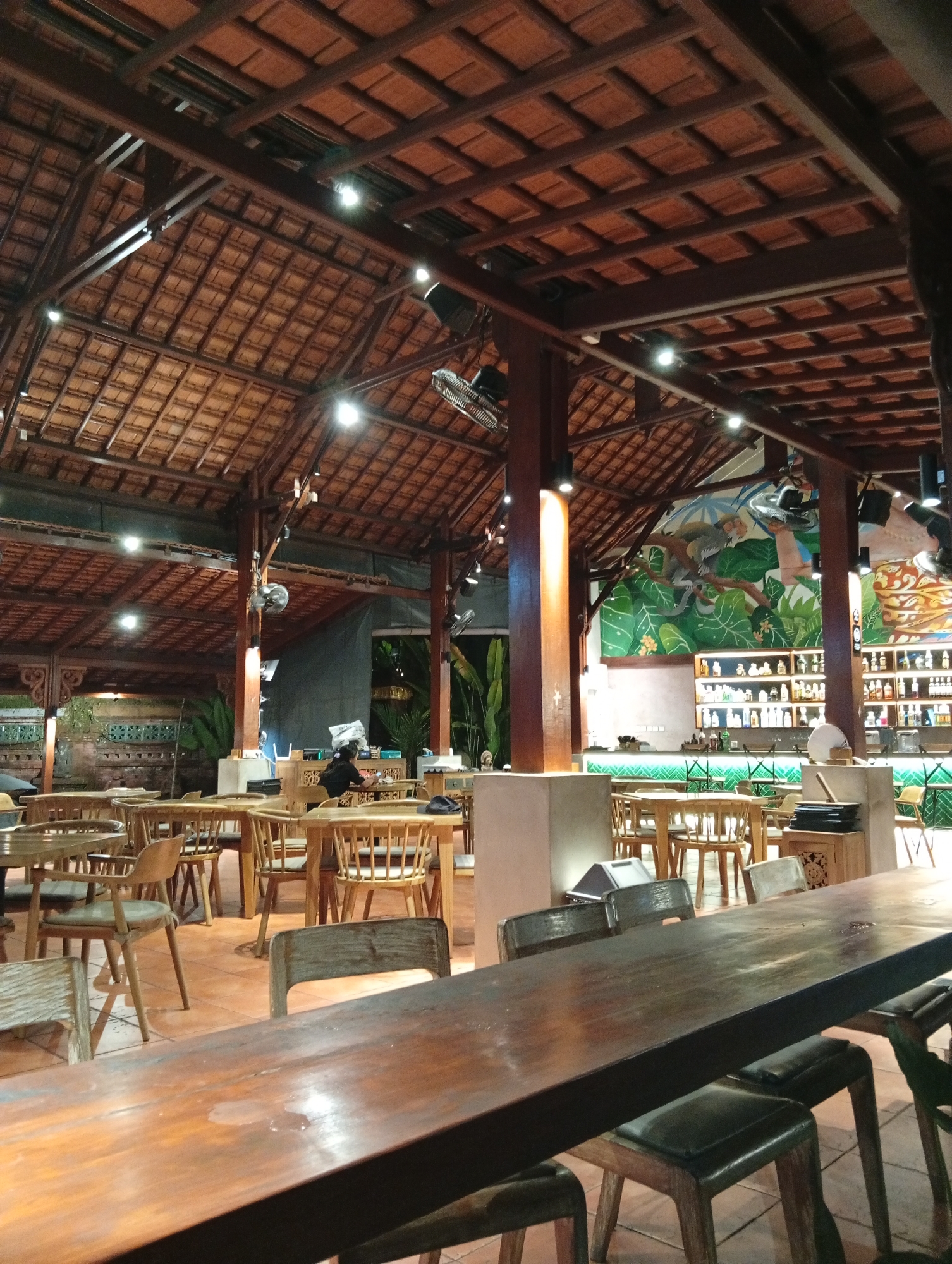 The image depicts the interior of a restaurant with a rustic, open-air design, likely in Ubud District, Indonesia, characterized by a high, tiled roof supported by wooden beams and pillars. The lighting is warm and ambient, emanating from spotlights and decorative lamps. The foreground features a long, polished wooden table with dark-seated chairs, suggesting a communal dining area.
In the midground, numerous wooden tables and chairs are arranged throughout the space, some appearing to be set for dining. A few patrons are visible: one individual is seated at a table in the middle distance, facing away from the camera, and another person appears to be working behind a counter on the right side of the image.
The background showcases a vibrant mural depicting monkeys and lush greenery, adding a tropical motif to the decor. Shelves stocked with bottles, likely beverages, are visible behind a bar area. The overall impression is a relaxed and inviting atmosphere within a tropical setting.