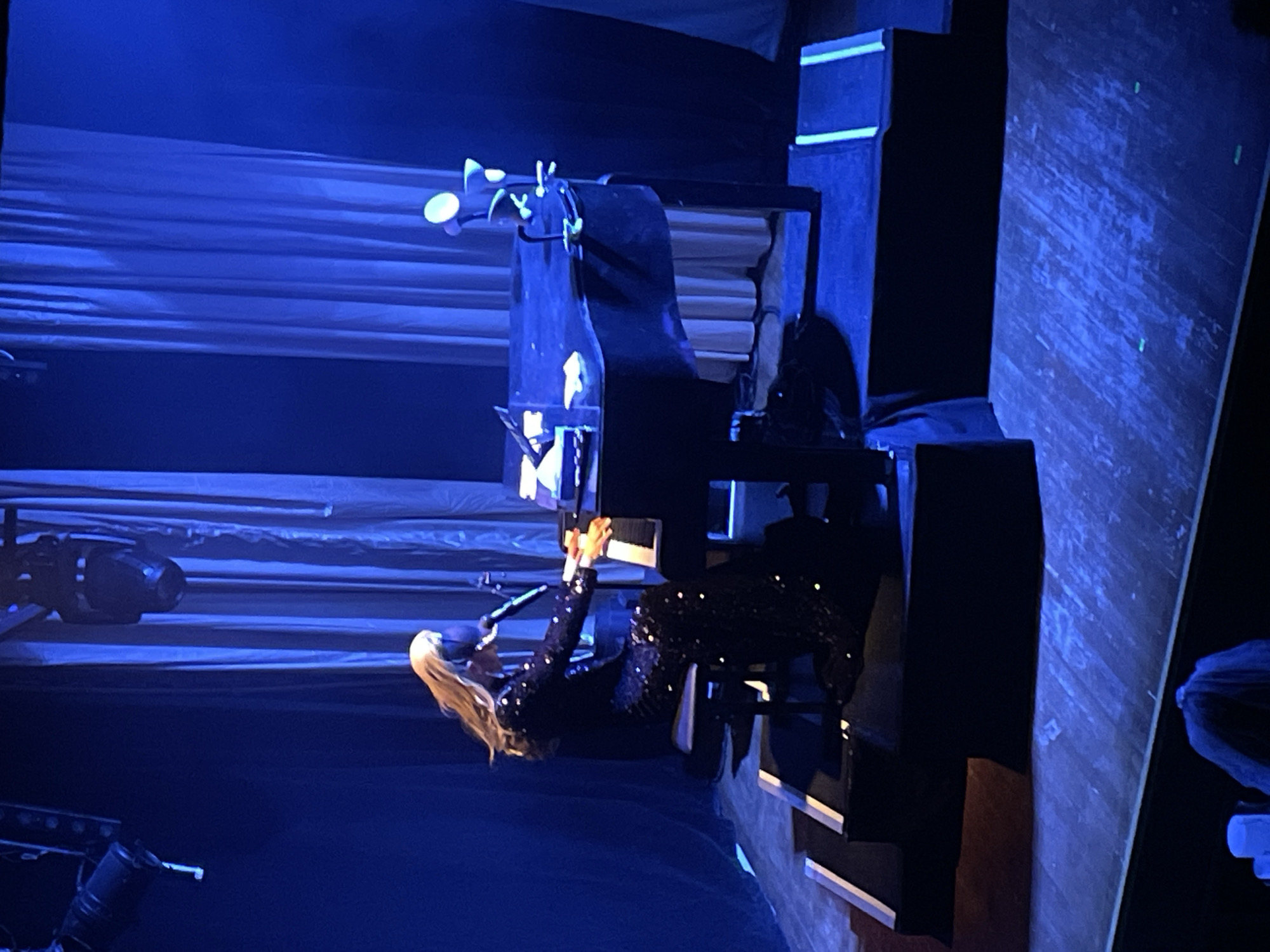 The image shows a woman playing a grand piano on a dimly lit stage. She is wearing a sparkly black gown and has long blonde hair. The stage is decorated with blue curtains and spotlights. The floor of the stage appears to be made of wood. The audience is not visible in the image. The setting is a performance venue, possibly a concert hall or theater. There is no indication that the scene is in Paris, France.