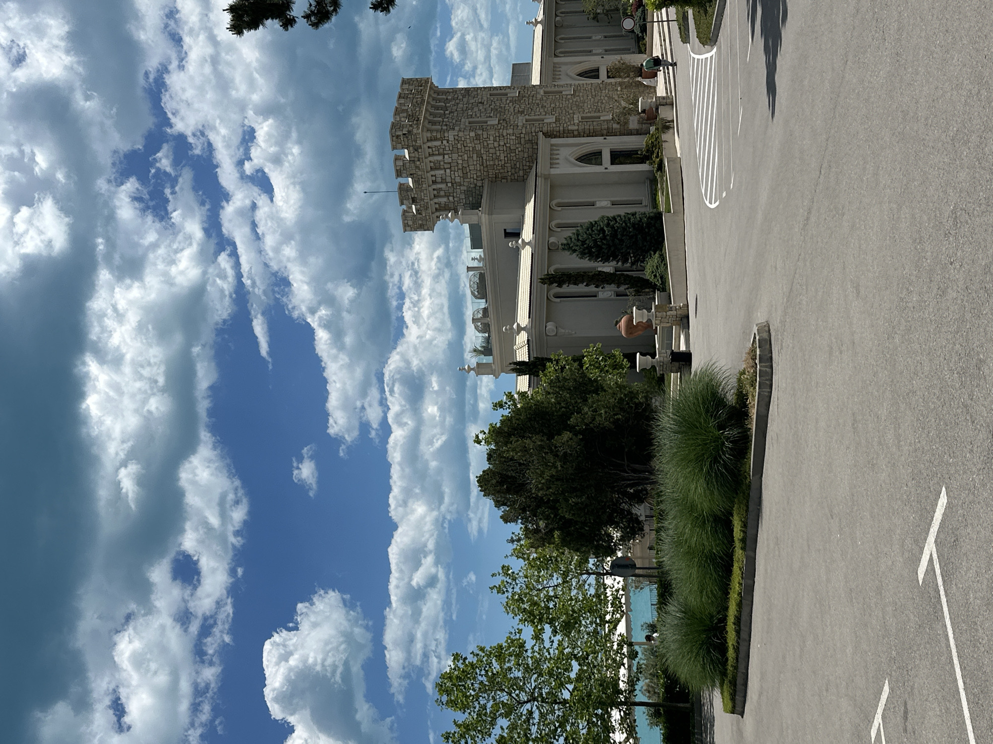 The image shows a building with a stone tower in Varna, Bulgaria, located next to a parking lot. The building is white with several windows and architectural details. Next to the building is a paved parking lot with white lines marking parking spaces. Adjacent to the parking lot are green trees and bushes. The sky is blue with puffy white clouds.