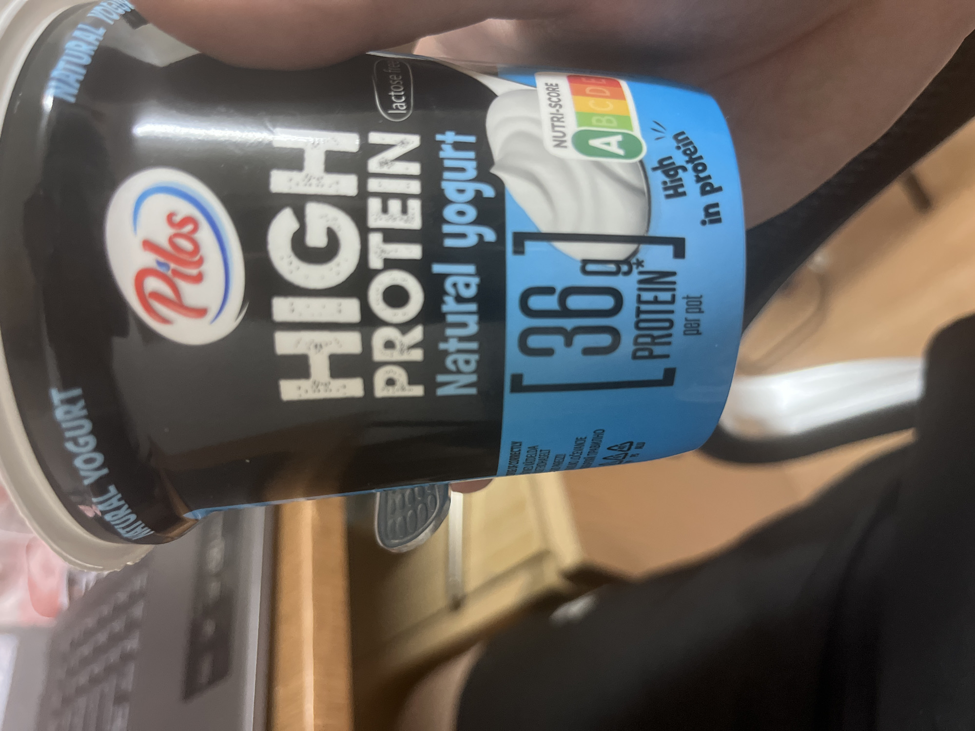 An overhead, partially rotated shot shows a hand holding a black yogurt pot, emphasizing its label. The product is a "Pilos HIGH PROTEIN Natural yogurt" and is explicitly marked "lactose free." A prominent blue section of the label highlights "36g PROTEIN per pot" and the claim "High in protein." The Nutri-score A is clearly displayed, indicating a favorable nutritional profile. Other visible text includes "NATURAL YOGURT" repeated on different parts of the packaging, and smaller multi-language instructions like "DLOČEVANJE" and "ПОРОЙ ПРАВИЛНО" (likely related to disposal in various languages, including Bulgarian given the context). The background is blurry, revealing wooden surfaces, suggesting an indoor setting like a desk or table, with glimpses of what might be office equipment or furniture. A black fabric, possibly clothing, is also visible in the background. The image focuses on the product and its nutritional information.