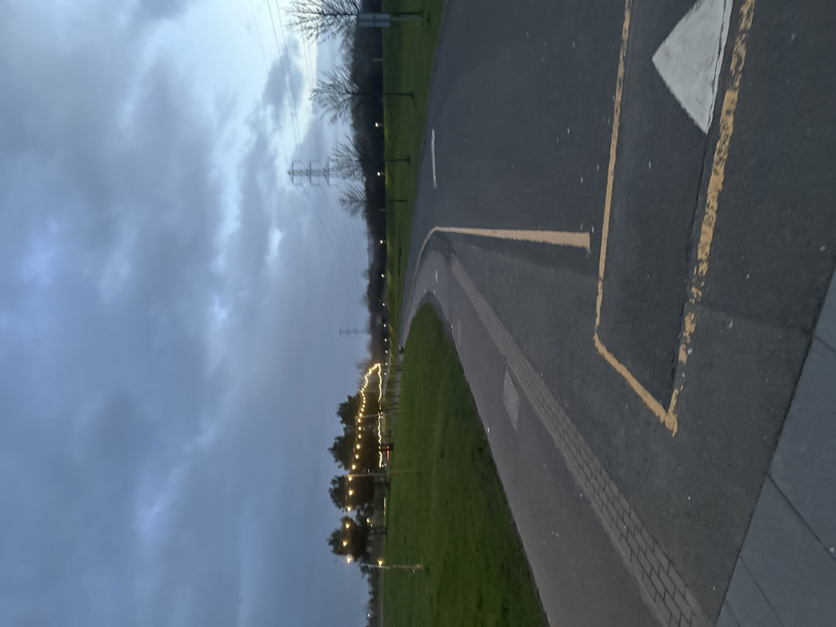 The image captures an outdoor park or recreational area in Falkirk, United Kingdom, consistent with the pathways and landscape of The Helix Park, during dusk or early evening under an overcast sky. No individuals are present.
In the foreground, a wide asphalt path or roadway curves from the lower right towards the mid-ground left. It features a continuous yellow line marking its center and a yellow rectangular box painted on the surface, within which an inverted white triangle (likely a yield or directional arrow) is visible. Adjacent and to the left, a separate, narrower paved pathway, partially surfaced with reddish brick pavers and partially asphalt, parallels the main path before curving left into the distance. Both paths are bordered by expansive, manicured green grass verges.
Along the left grassy verge, a string of warm yellow decorative lights is strung between a series of upright poles or slender trees, extending in an arc into the mid-ground. Further along this side, a cluster of trees with visible foliage is present. To the right of the main path, a sparse line of bare-branched deciduous trees stands. In the distant background, several tall metal lattice transmission towers (pylons) are silhouetted against the dark, cloudy sky, indicating a power line route. Additional individual streetlights or park lights are visible illuminating the path further in the distance. The sky is uniformly grey-blue with substantial cloud cover.
