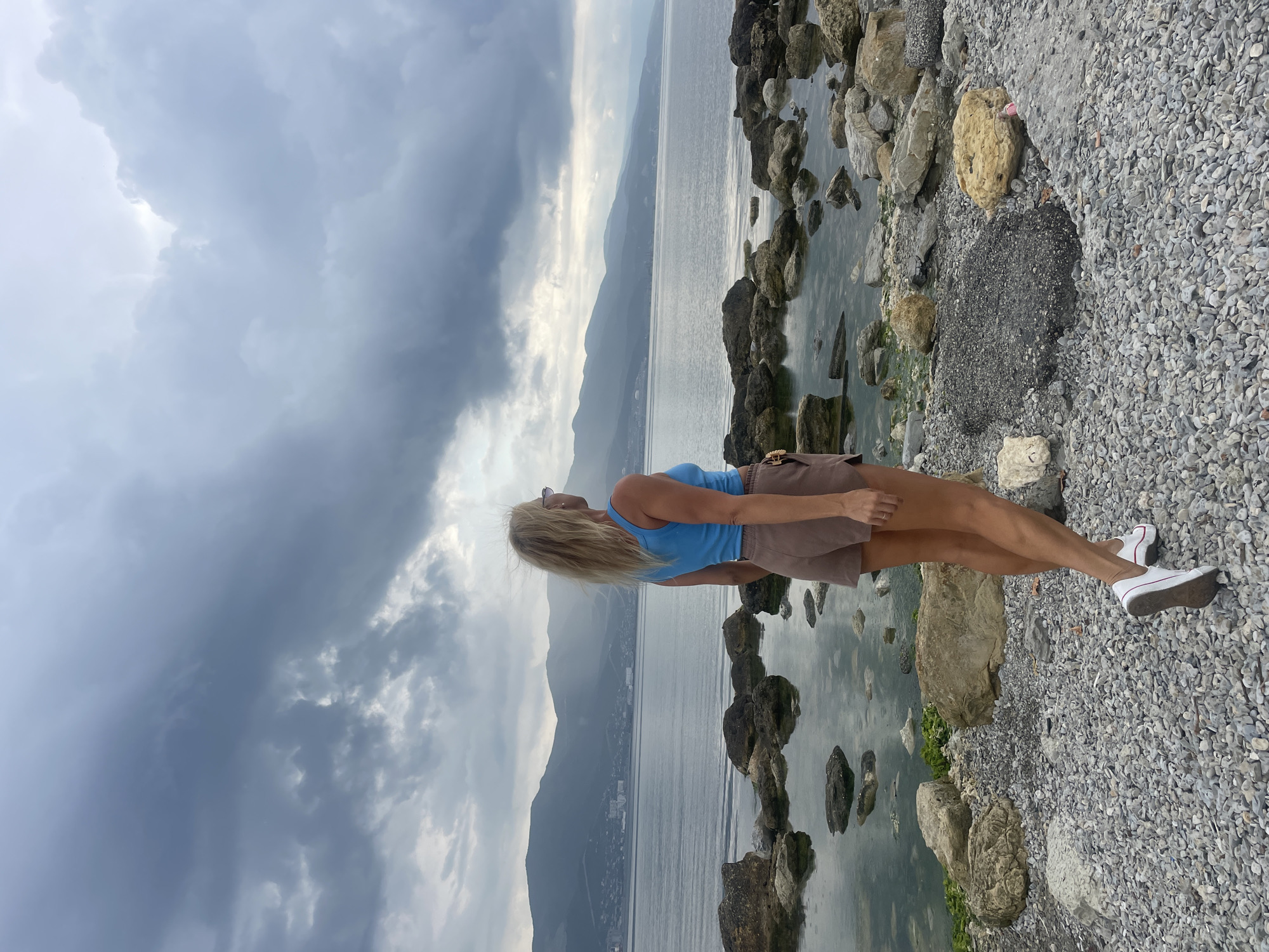 A blonde woman wearing a blue tank top and brown shorts stands on a rocky shoreline, facing away from the camera and looking out at the sea. The water is calm, and in the distance, there are mountains and a city skyline, possibly Novorossiysk, Russia. The sky is overcast with dark, dramatic clouds. The ground is covered in small pebbles and larger rocks.