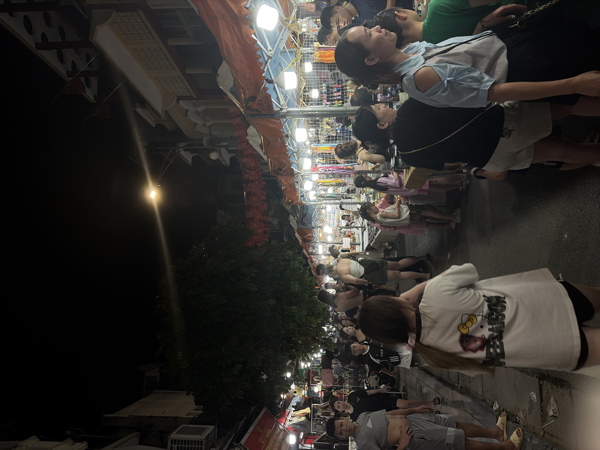 This is a nighttime scene of a bustling street market, likely in Hanoi, Vietnam, as suggested by the presence of Vietnamese flags and the general atmosphere of a night market. A diverse group of people, including men, women, and children, are walking through the market, browsing goods displayed at various stalls. The stalls are illuminated by string lights and overhead lamps, creating a vibrant and lively ambiance. Many stalls appear to be selling accessories like jewelry, keychains, and other small trinkets. Some stalls also display clothing items. The architecture visible in the background suggests a mix of traditional and modern buildings. The overall scene conveys a sense of community and activity, typical of a local market at night.